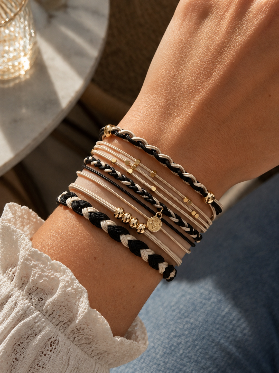 Lot de 5 élastiques à cheveux et bracelets 2-en-1 noirs, crème et dorés avec tresses bicolores, perles dorées et breloque médaillon, portés au poignet.