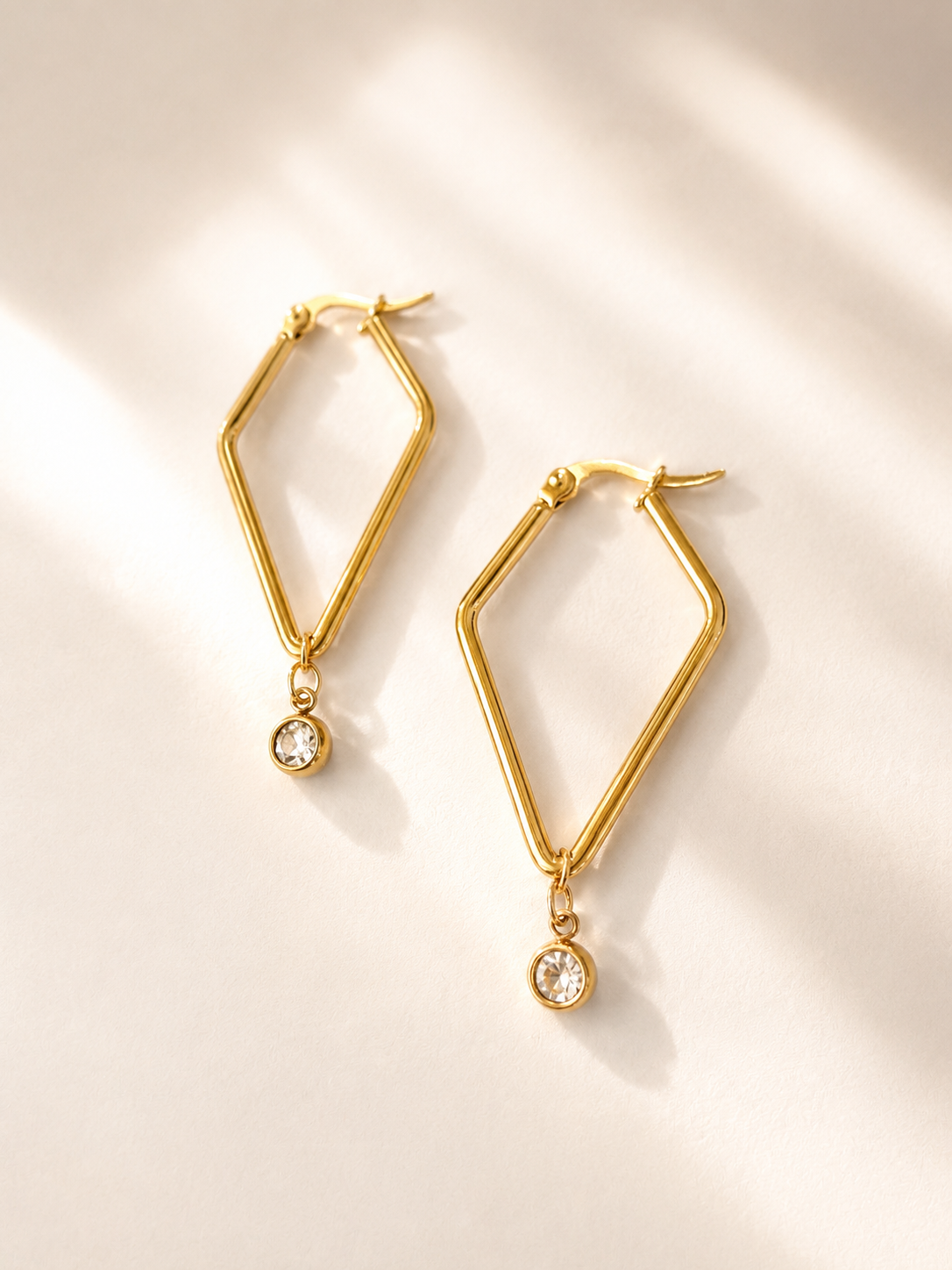 Boucles d’oreilles Vivienne en acier inoxydable doré de forme géométrique avec cristal suspendu sur fond blanc. Par Tokade