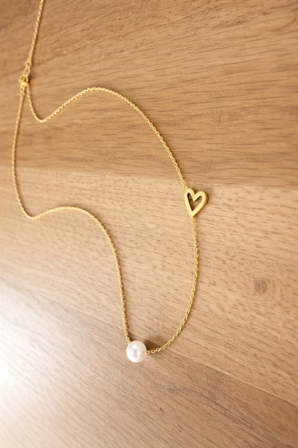 Vue rapprochée du collier Joan en acier inoxydable doré. La finesse de la chaîne, la perle centrale et le cœur minimaliste sont mis en valeur sur un fond naturel en bois clair.