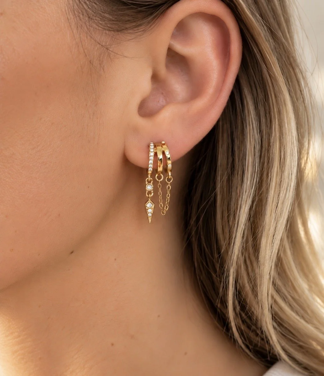 Boucles d’oreilles Jenna dorées à trois anneaux ornés de pierres brillantes et de chaînes pendantes, portées sur une oreille féminine.