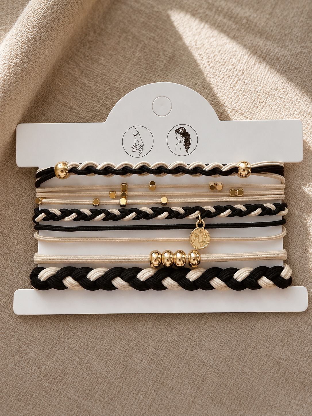 Lot de 5 élastiques à cheveux et bracelets 2-en-1 noirs, crème et dorés avec tresses bicolores, perles dorées et breloque médaillon, portés au poignet.