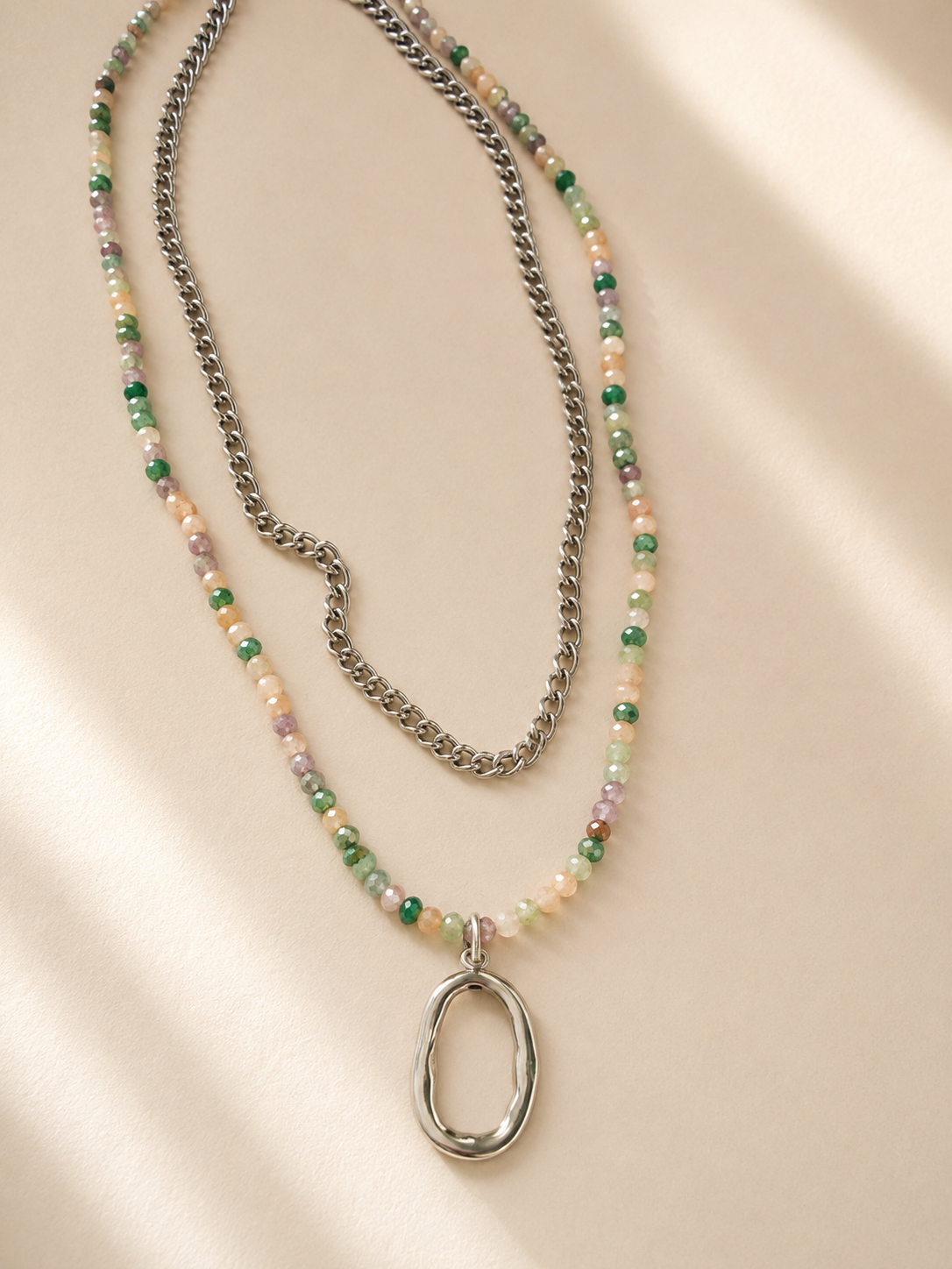 Collier Saya double rang en acier inoxydable argenté avec pierres naturelles multicolores et pendentif organique sur fond crème. - Tokade