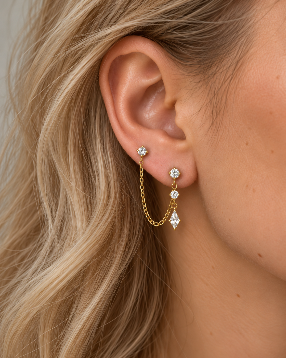 Boucles d’oreilles Akina dorées portées sur deux trous d’une oreille féminine, avec chaîne tombante et pierres brillantes.