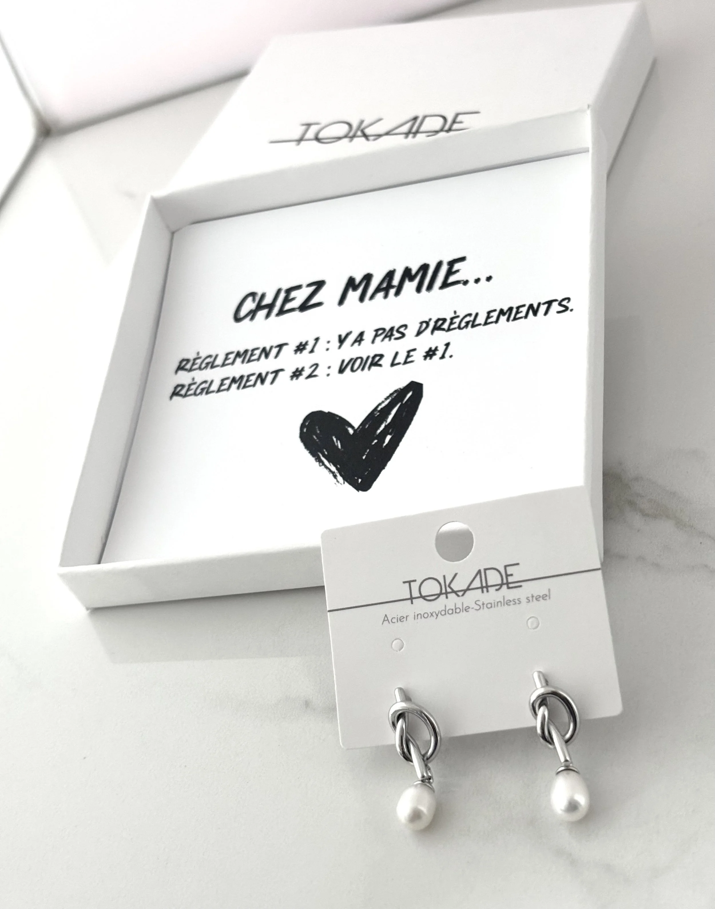 Boîte cadeau Tokade avec le message humoristique « Chez mamie… y’a pas d’règlements » et boucles d’oreilles Selya en acier inoxydable argenté avec perle blanche.