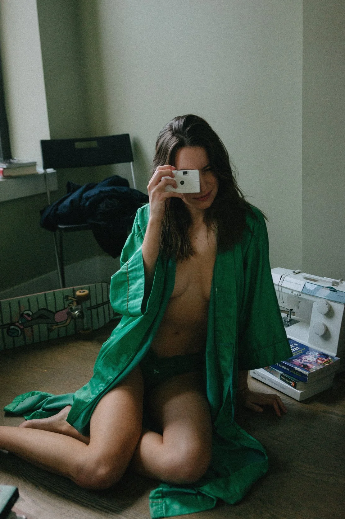 A woman is kneeling on the floor, taking a mirror selfie with her phone. She is wearing a green robe, black underwear, and is topless. The room has a skateboard, books, a sewing machine, and other personal items.