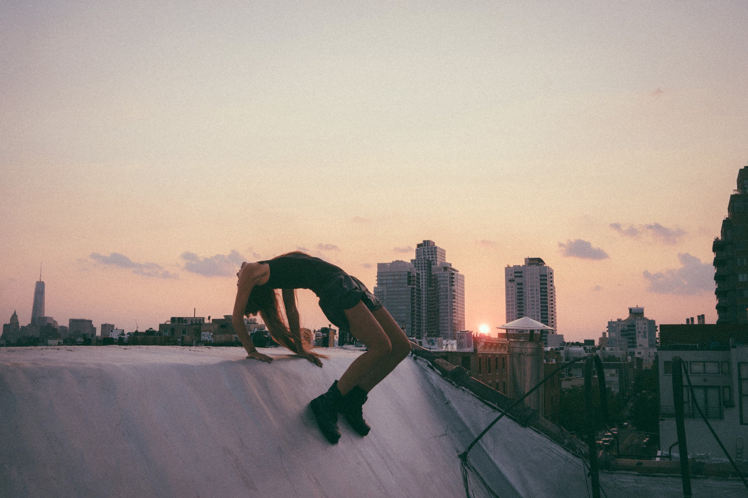 A person with long hair wearing a black tank top and shorts performing a backbend yoga pose on a rooftop at sunset, with a city skyline in the background.