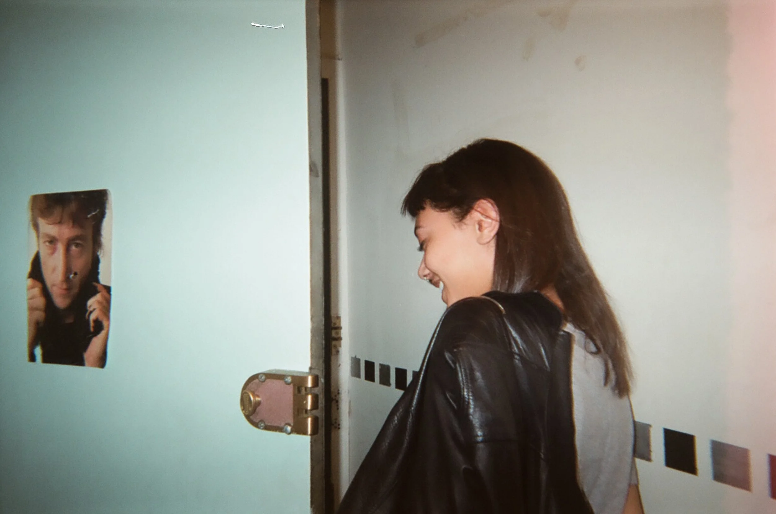 A woman with dark hair and a leather jacket standing in a hallway, smiling with her eyes closed, facing a closed door with a photo of a man on it.