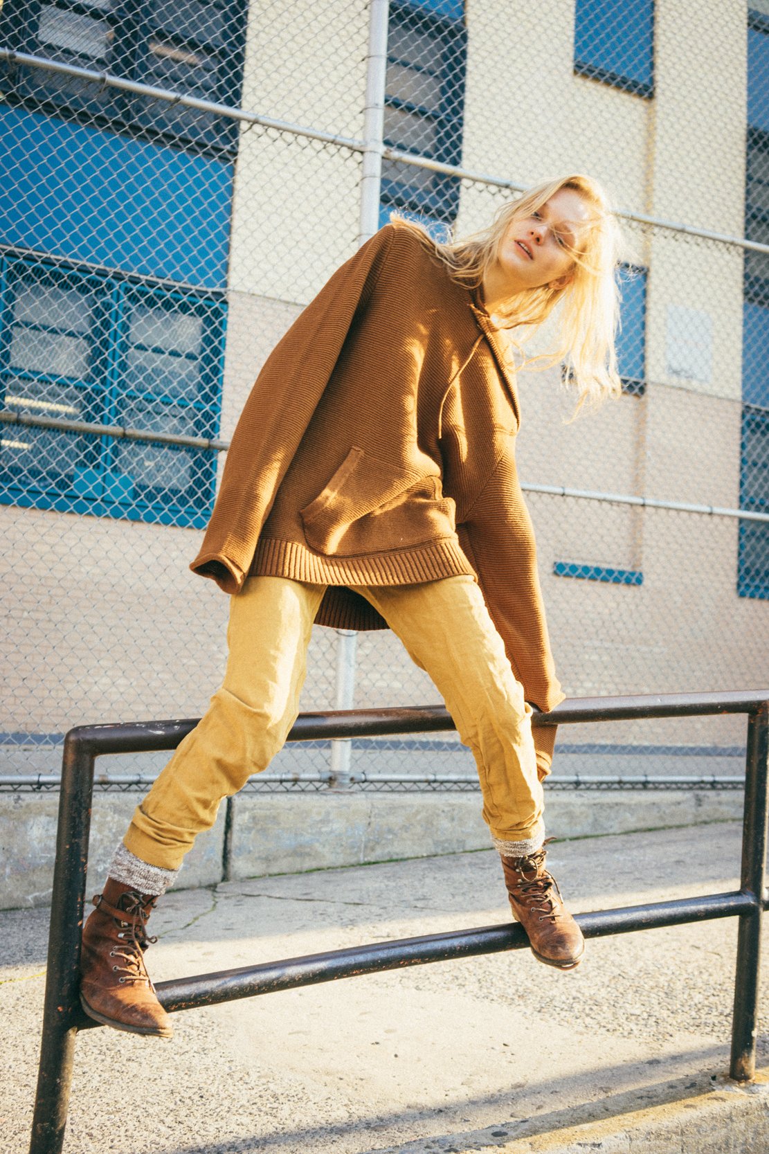 A young woman with blonde hair poses on a black metal rail outdoors near a chain-link fence and a building with blue windows, dressed in a brown oversized hoodie, tan pants, and brown boots.