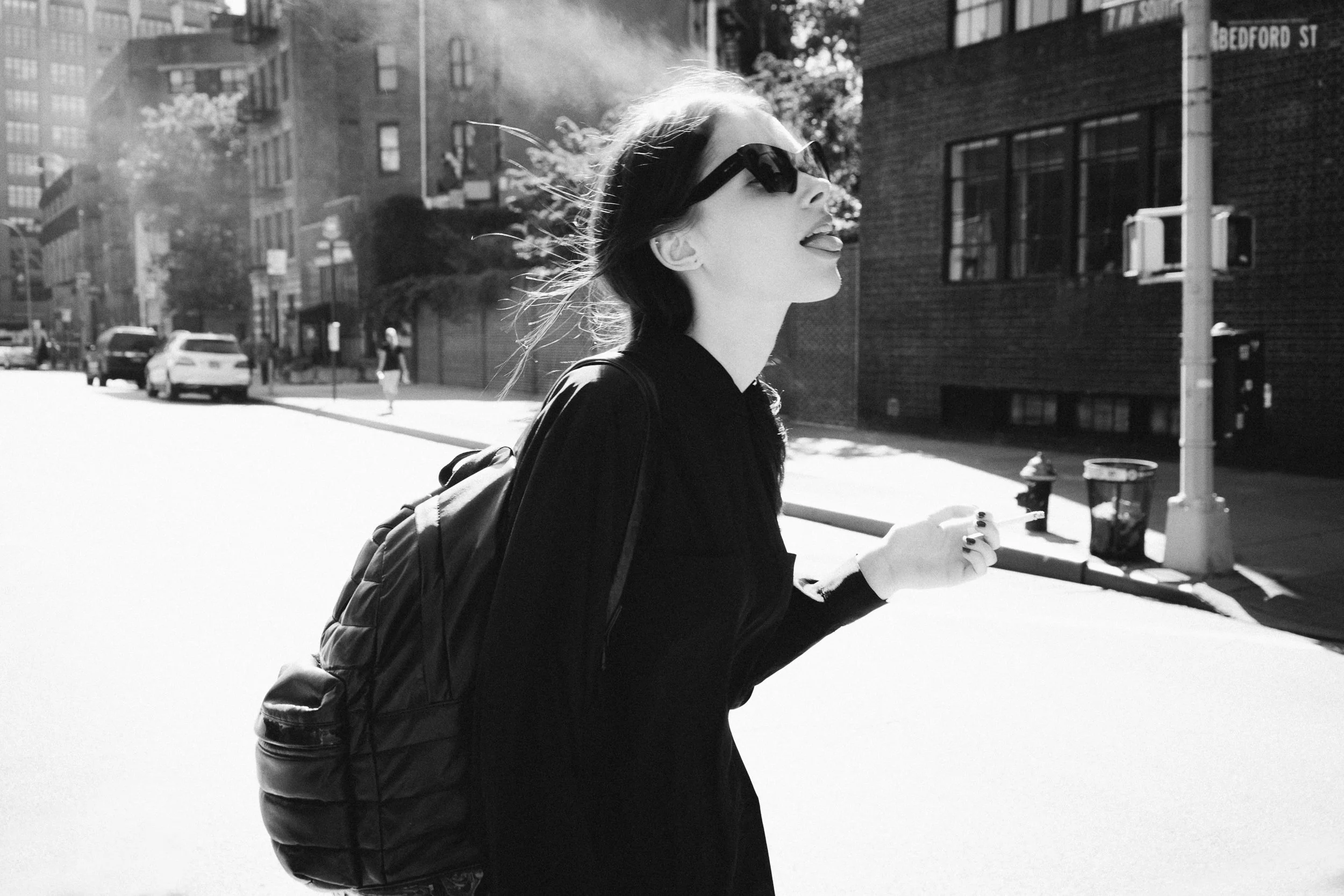 A woman with sunglasses and a backpack smiling and sticking out her tongue while walking on a city street.