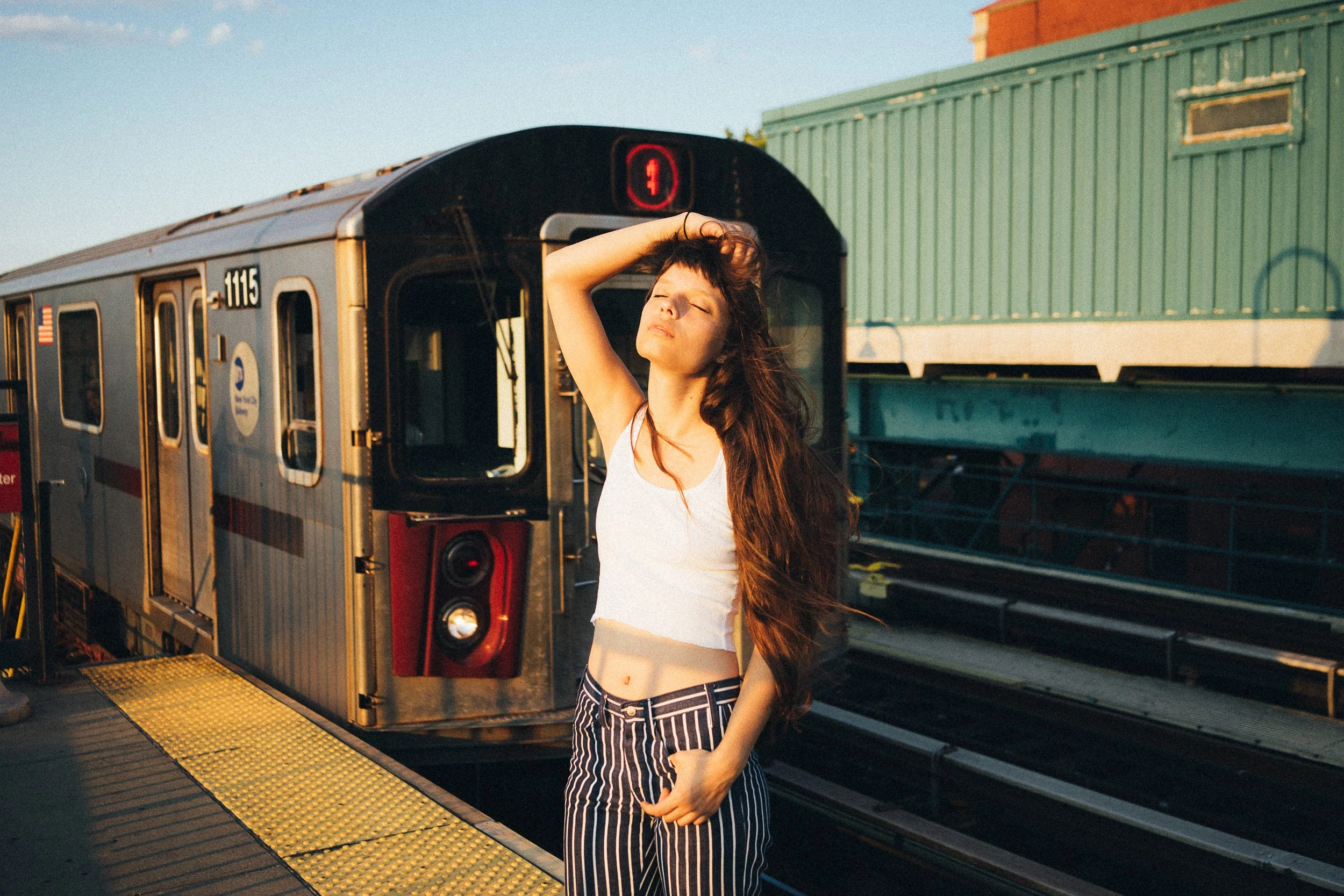 Young woman with long hair standing on a subway platform near an incoming train, appearing relaxed with eyes closed and one hand running through her hair.