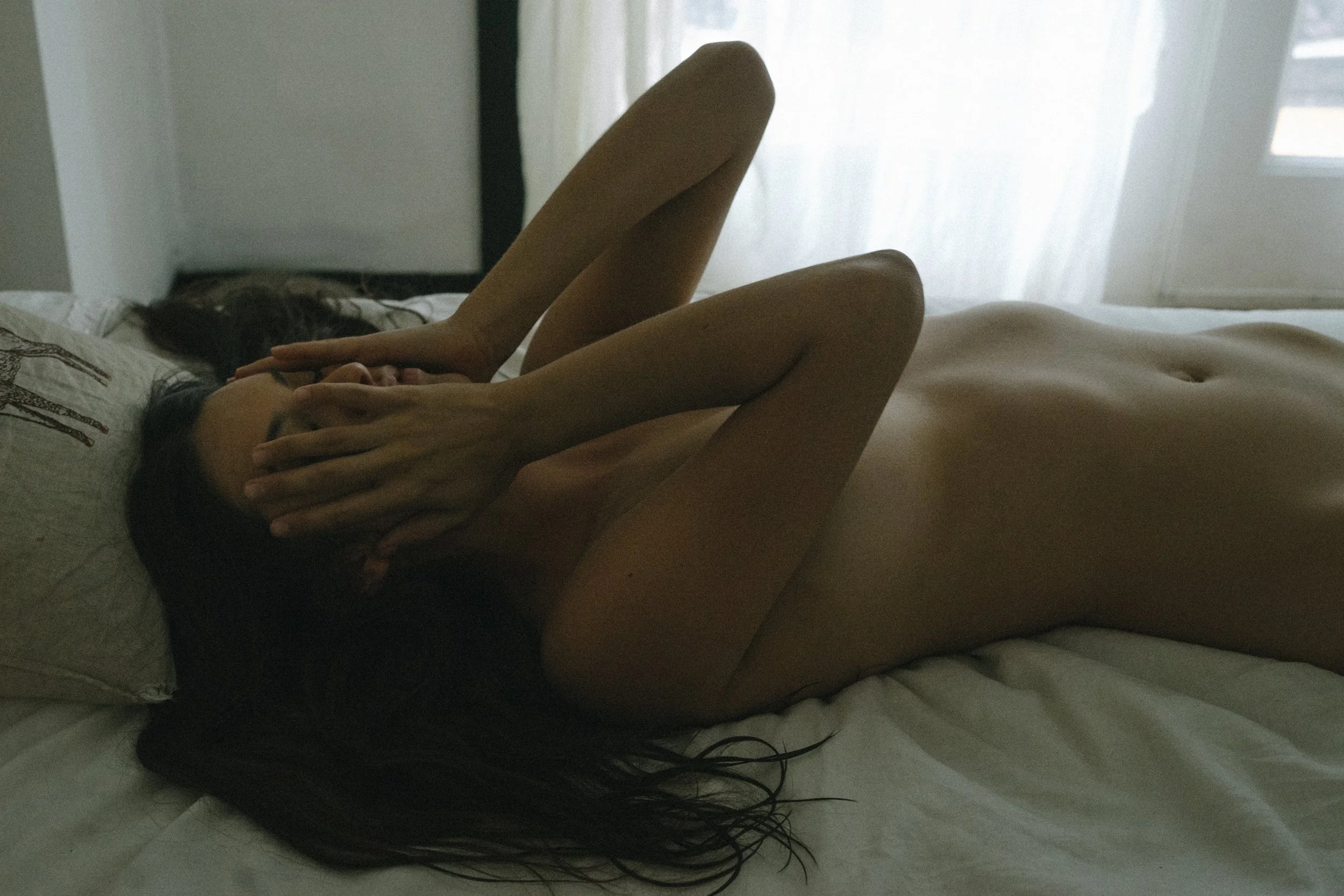 A woman lying on a bed with her hands covering her face, in a softly lit room with sheer curtains.