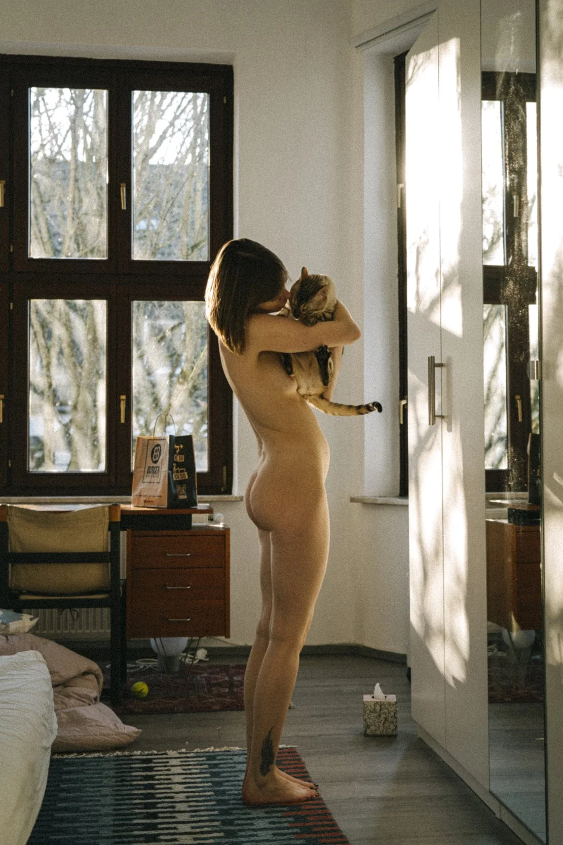 A naked woman holding a cat in a bedroom lit by natural sunlight.
