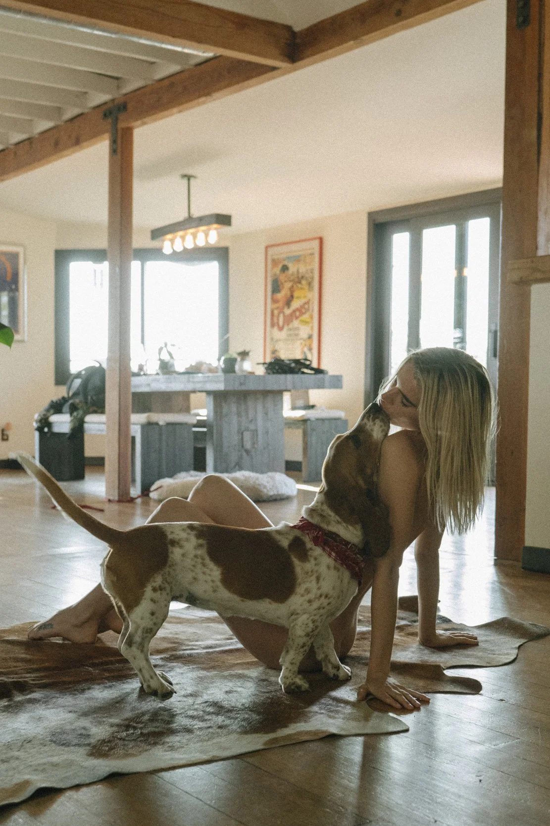 A woman and a dog in a cozy home interior. The woman is sitting on the floor, leaning on her hands, and the dog is nuzzling her face. Sunlight streams through large windows in the background, illuminating the room.