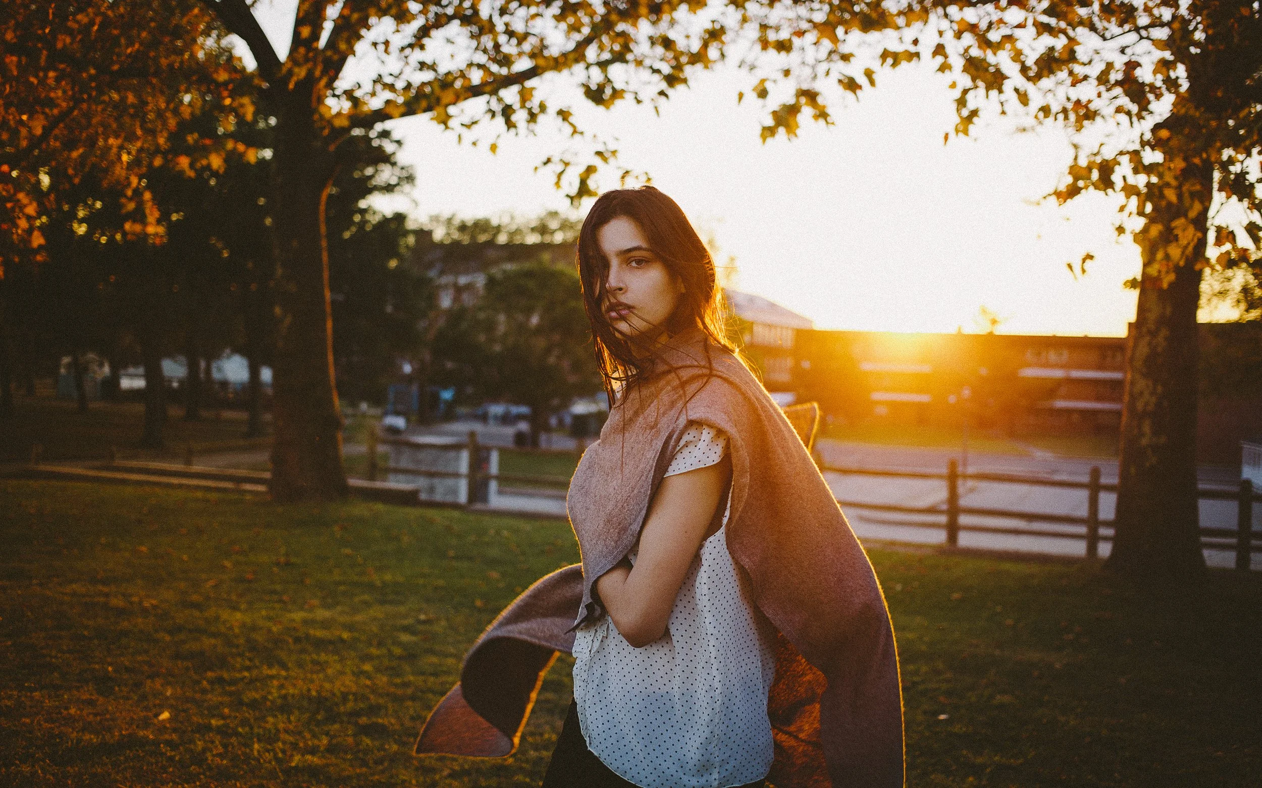 A young woman standing outdoors in a park during sunset, wearing a light blouse and a brown coat draped over her shoulders. She looks at the camera with a serious expression, with trees and a fence behind her.