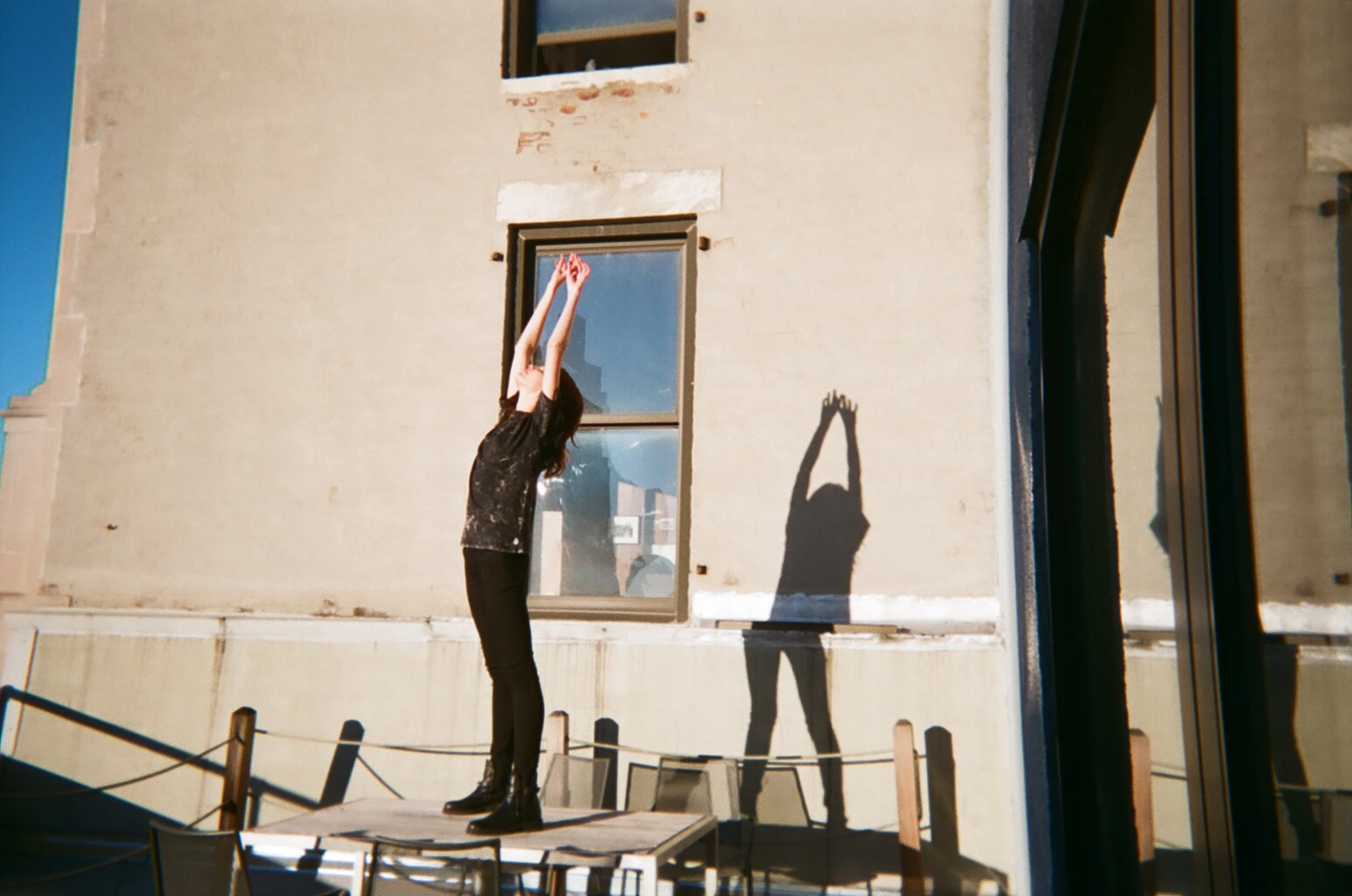 A woman in black standing on a table with her arms raised above her head, and her shadow cast on a building wall behind her.