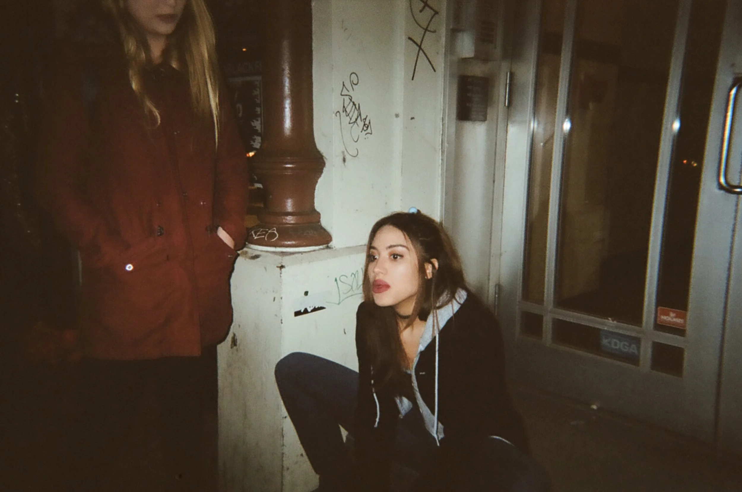 Two young women outside at night, one is standing in a red jacket with hands in pockets, the other is sitting on the ground with dark hair, wearing a black hoodie and observing something to the right.