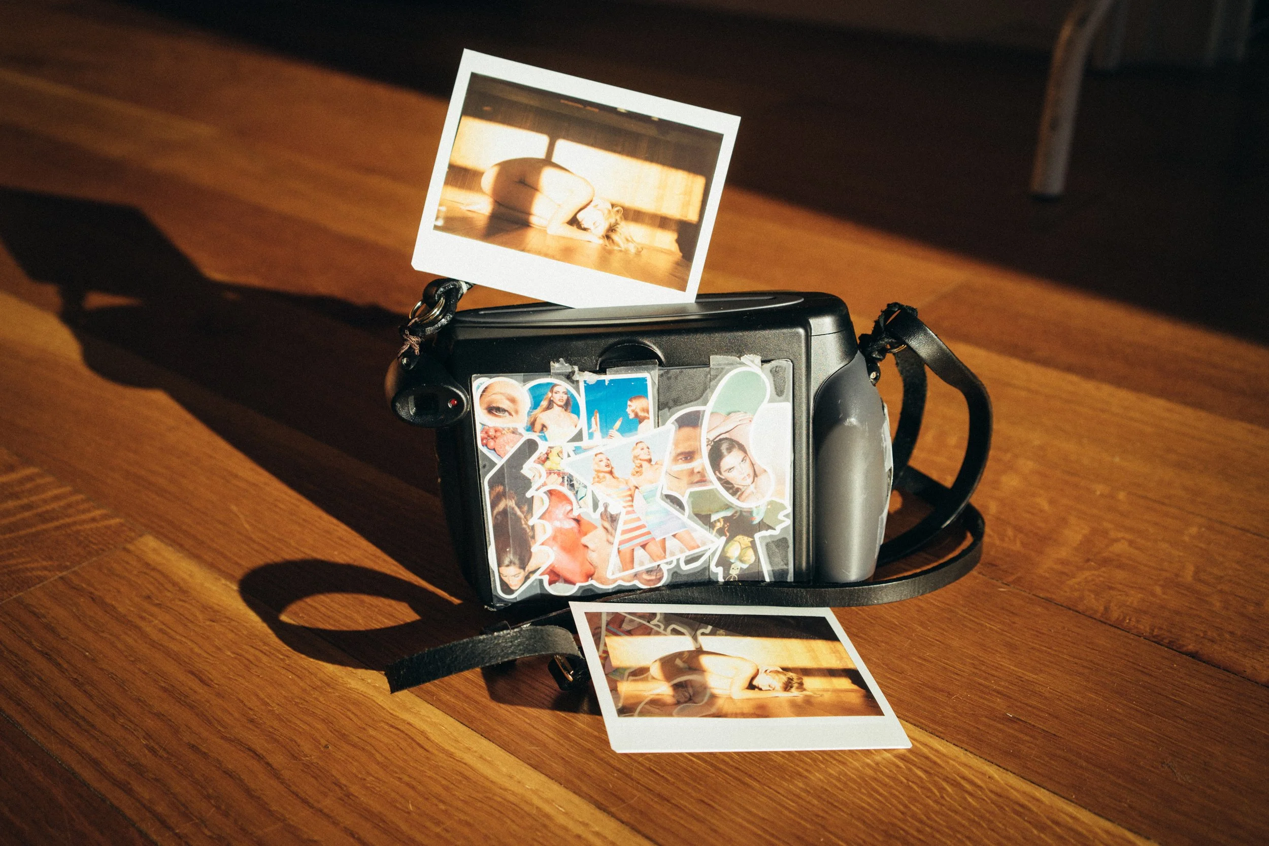 A black camera on a wooden surface with four Polaroid photos attached, showing images of women, a beach scene, and a person practicing yoga in sunlight.