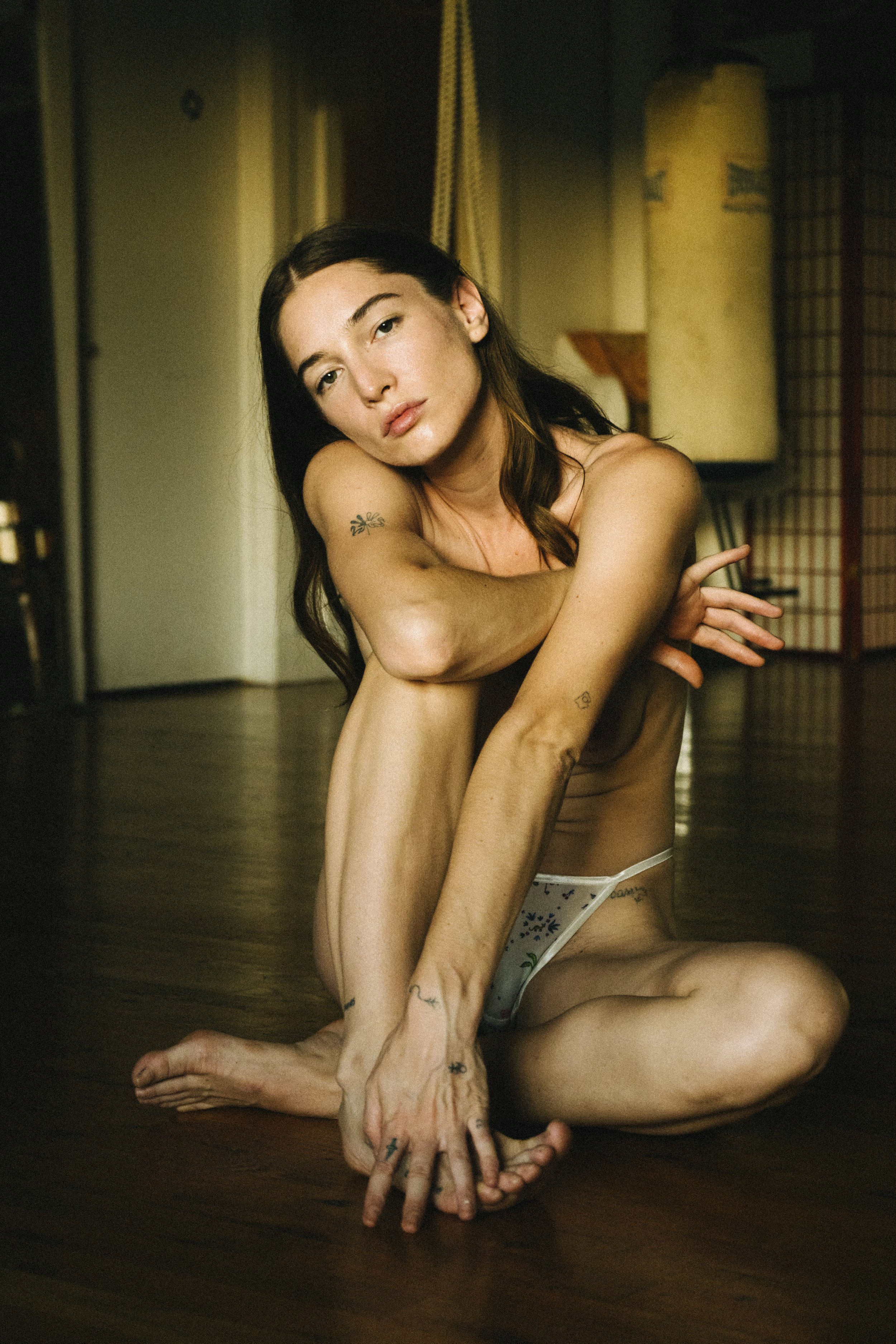 A woman with dark hair and tattoos sitting on a wooden floor, wearing only underwear, with her arms crossed over her knees and looking into the camera.