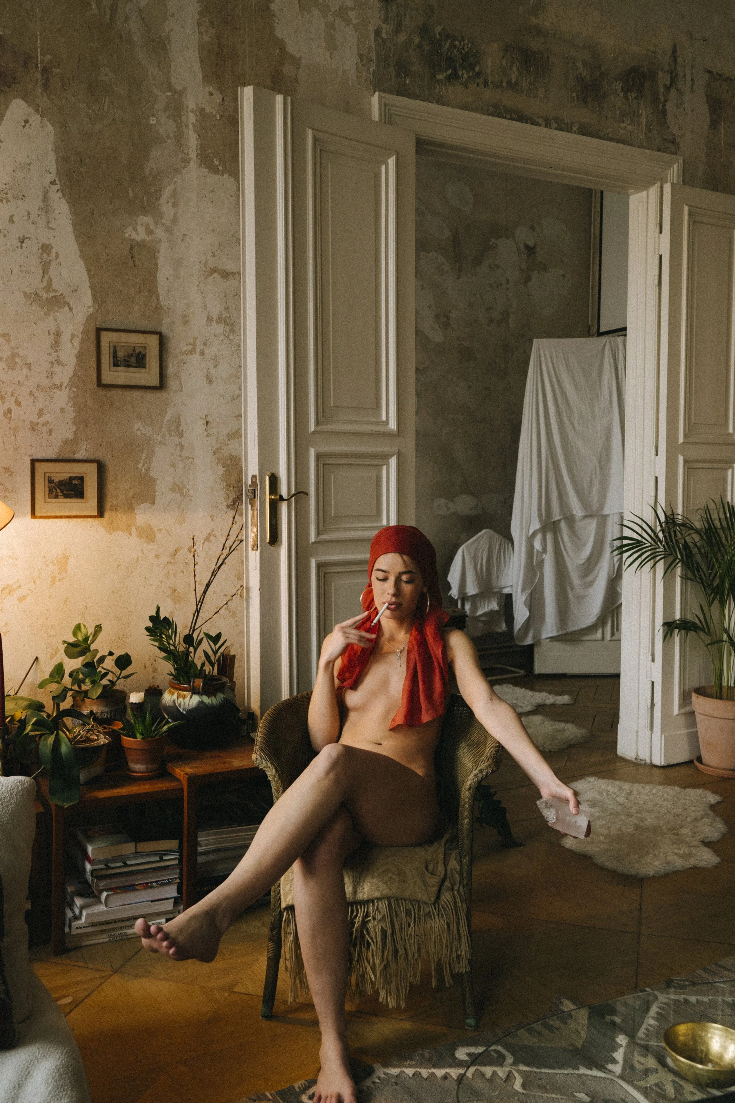 A woman with a red headscarf sitting nude in a vintage armchair in a room with peeling walls, plants, and a wooden floor.
