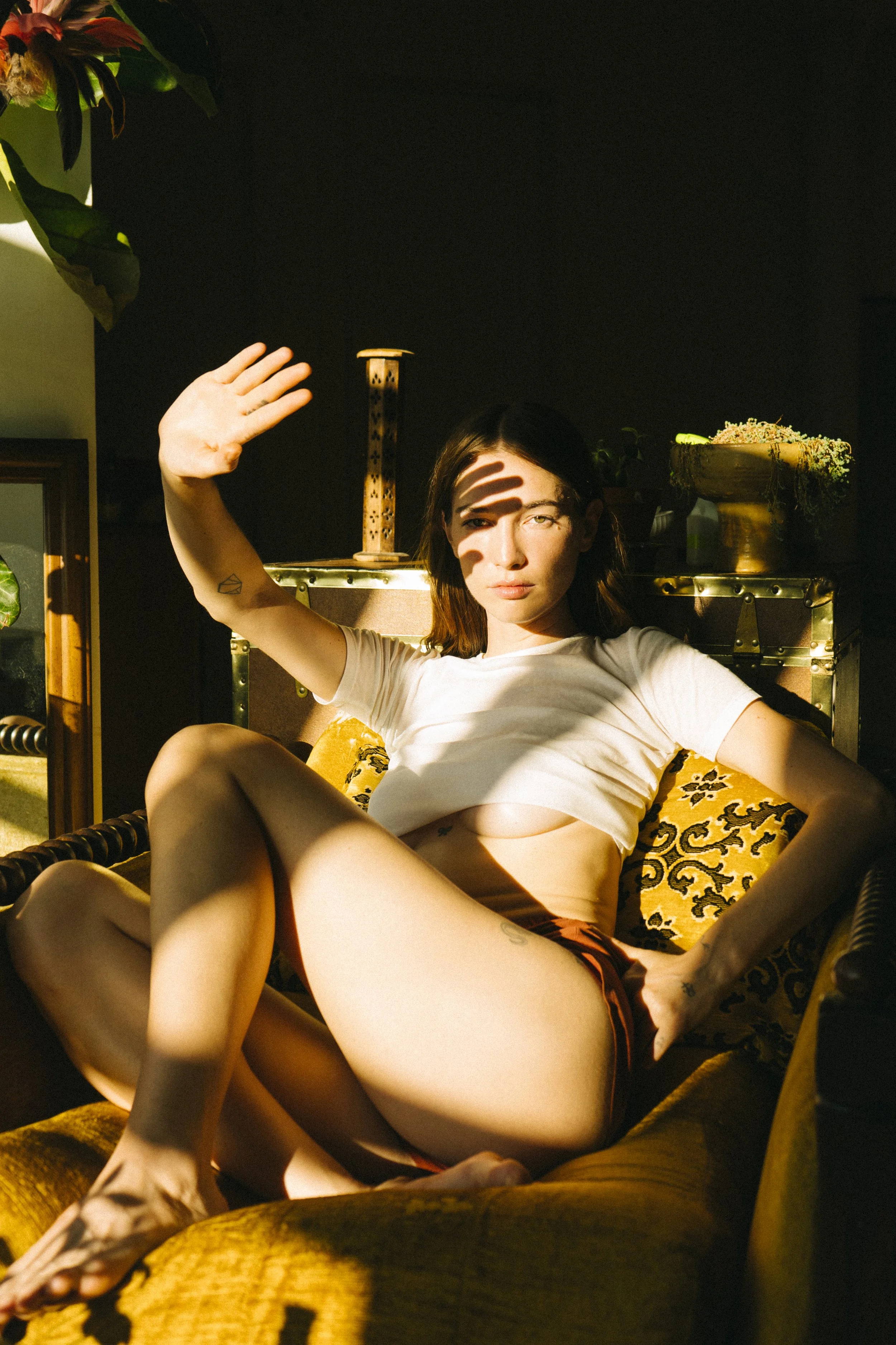 A woman sitting on a vintage yellow couch, raising her hand to shield her eyes from sunlight, with shadows cast across her face and body.