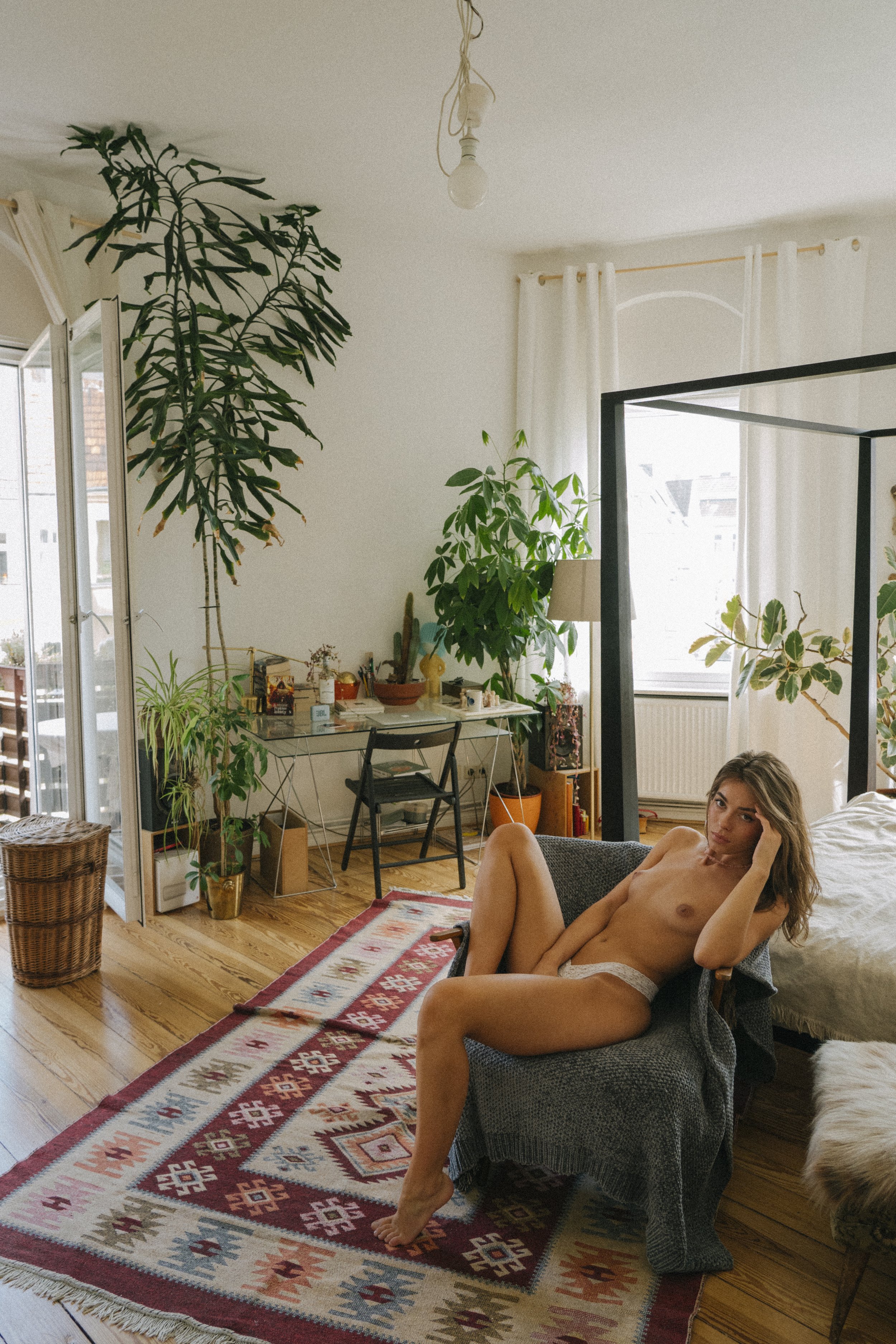 A woman is sitting nude on a gray armchair in a sunlit living room, surrounded by plants, a colorful patterned rug, and various furniture and decor items.