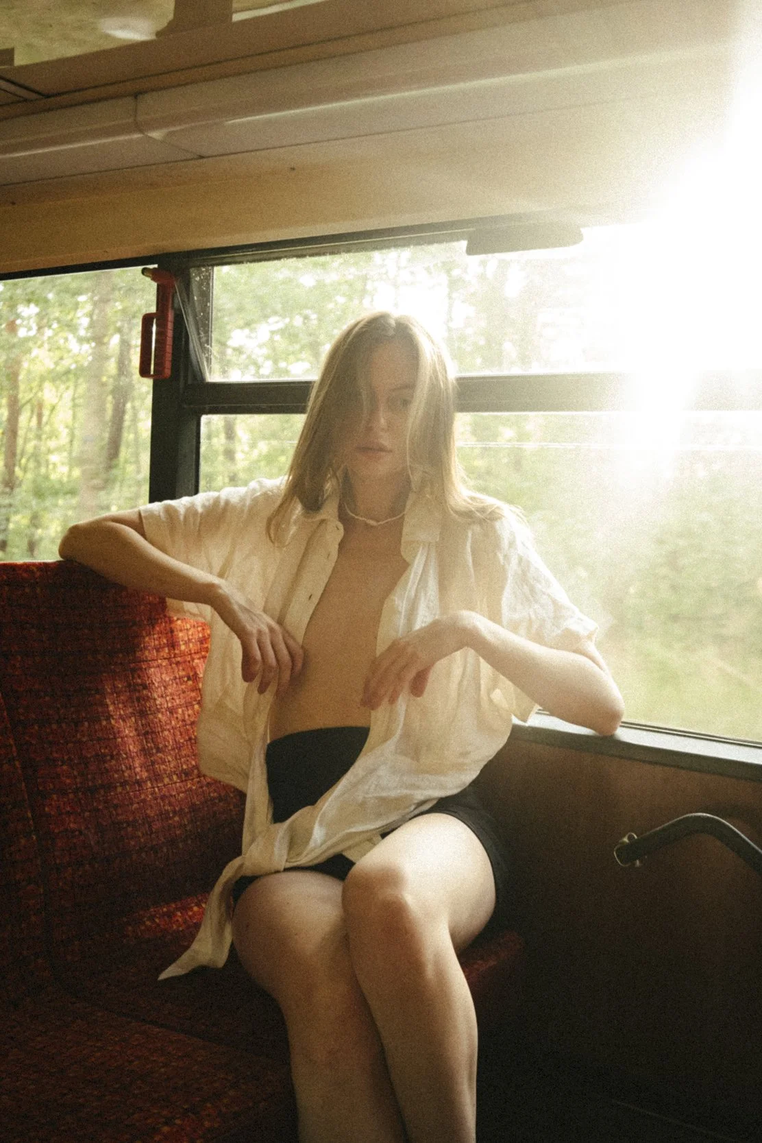 A young woman with blonde hair, wearing a loose beige shirt and black shorts, sitting on a red patterned seat inside a bus with a window showing a green, leafy outdoor scene, and sunlight streaming in.