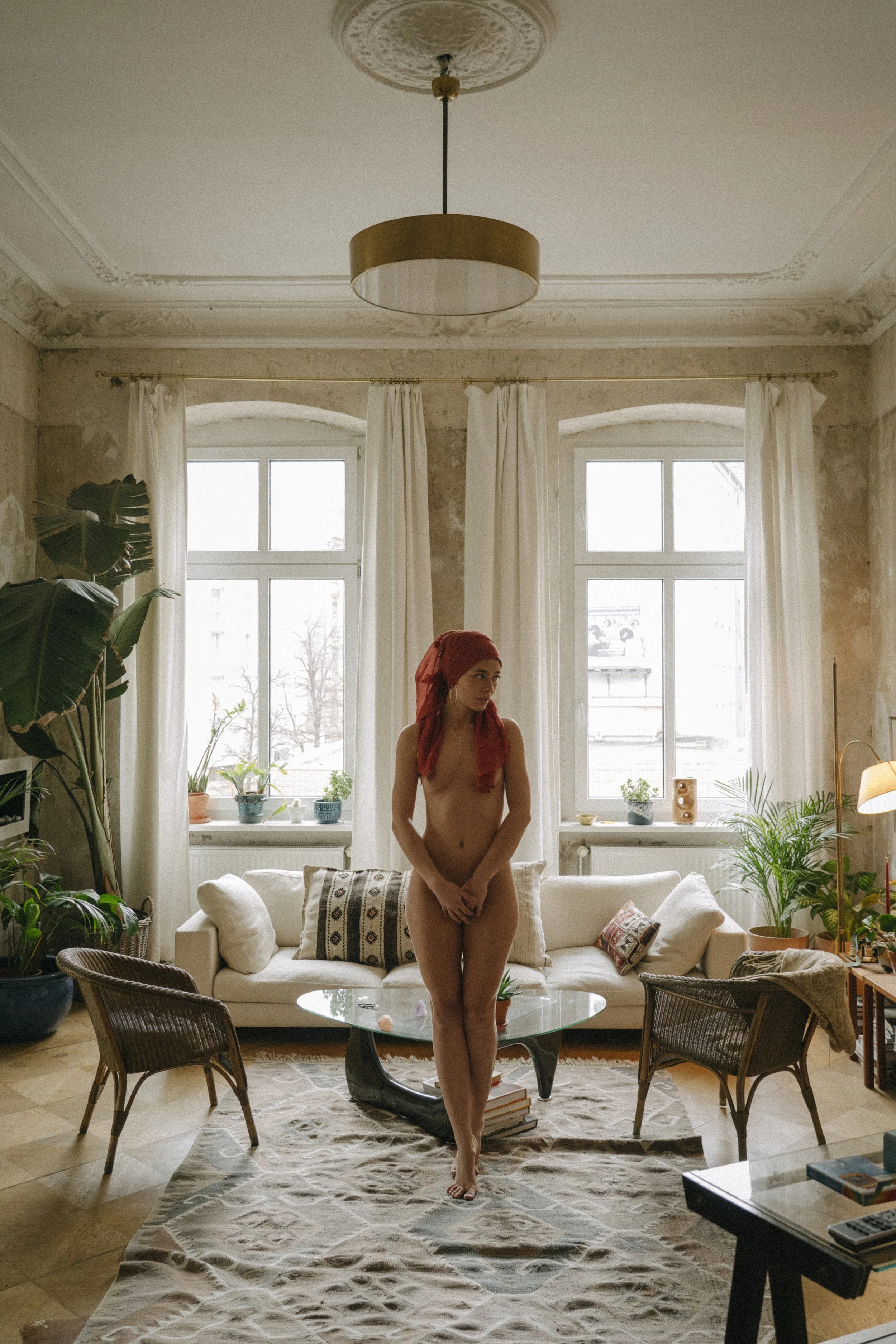 A woman standing nude in a living room with white walls, large windows, potted plants, a white sofa, and a patterned rug.