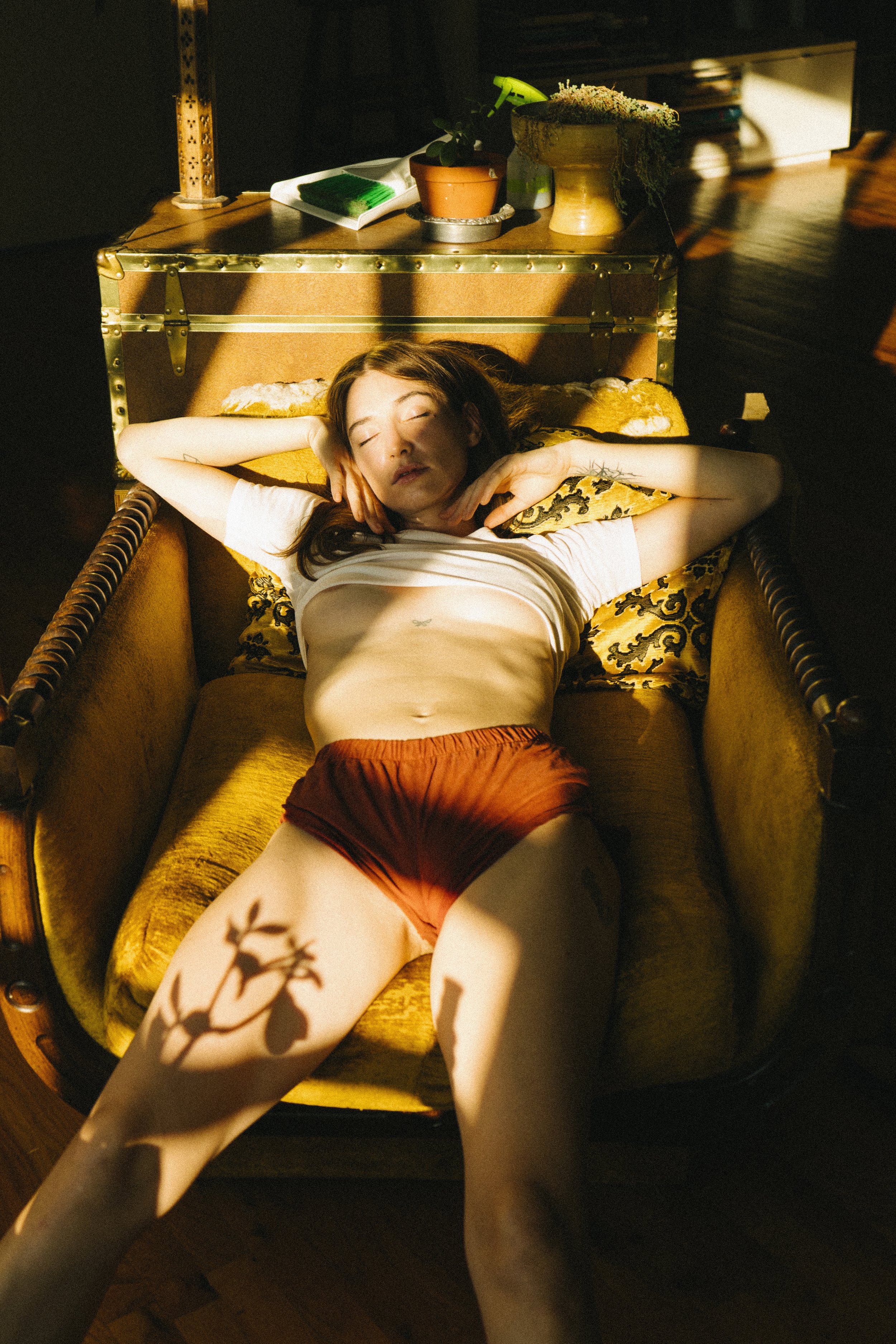 A woman with reddish-brown hair is lying on her back on a yellow velvet couch with her eyes closed, wearing a white t-shirt and red shorts. Sunlight casts a shadow of a plant's branches and leaves on her body.