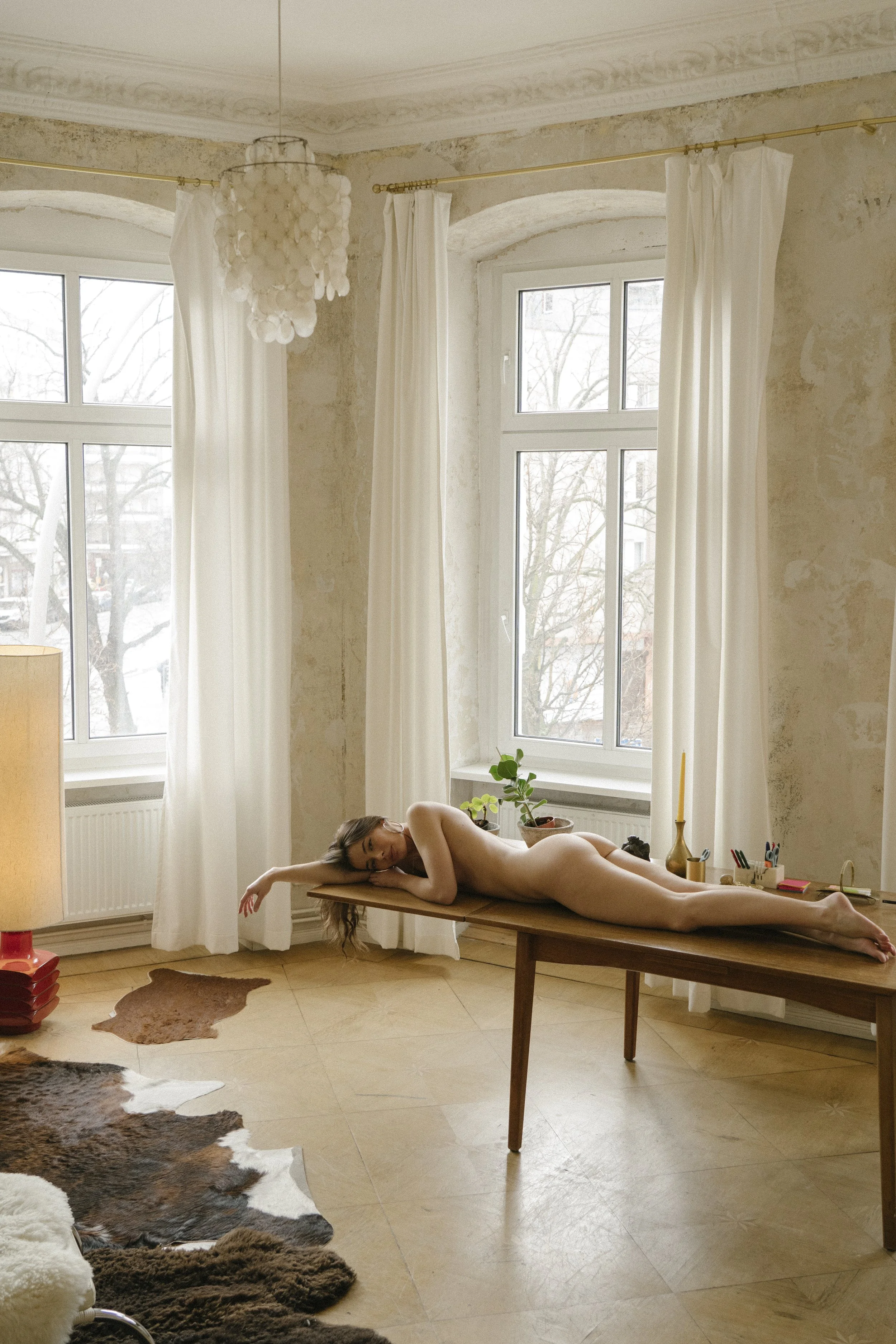 A nude woman lying on her side on a wooden table in a well-lit room with large windows, white curtains, hardwood floors, and a cowhide rug.