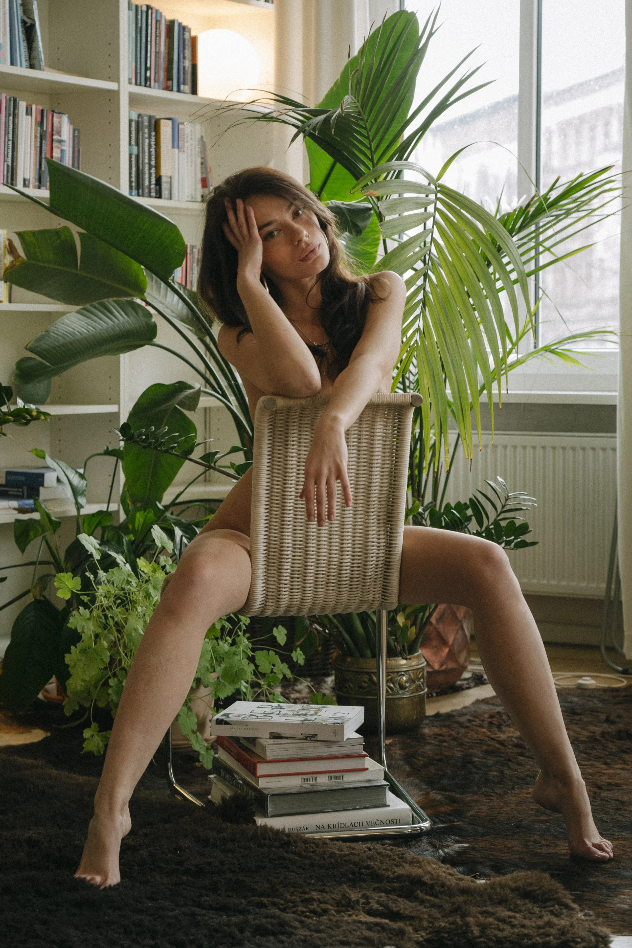 A woman sitting on a chair in a room filled with large green plants, with her legs spread apart and her hand on her forehead. There are books stacked beneath the chair and a bookshelf with more books in the background, with a window letting in natura