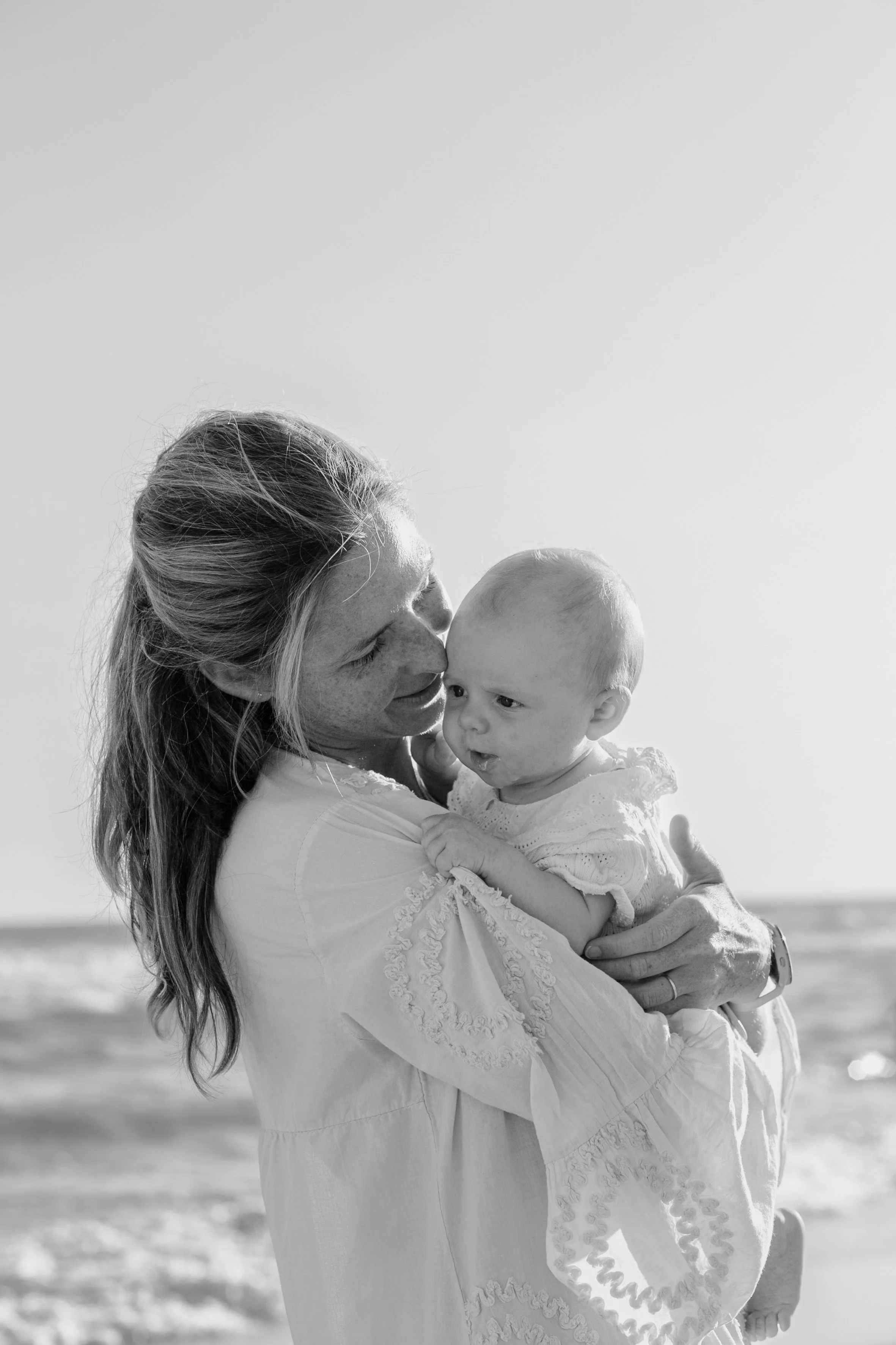 A woman holding a baby on a beach, both dressed in white, with the woman kissing the baby's forehead. The image is in black and white.