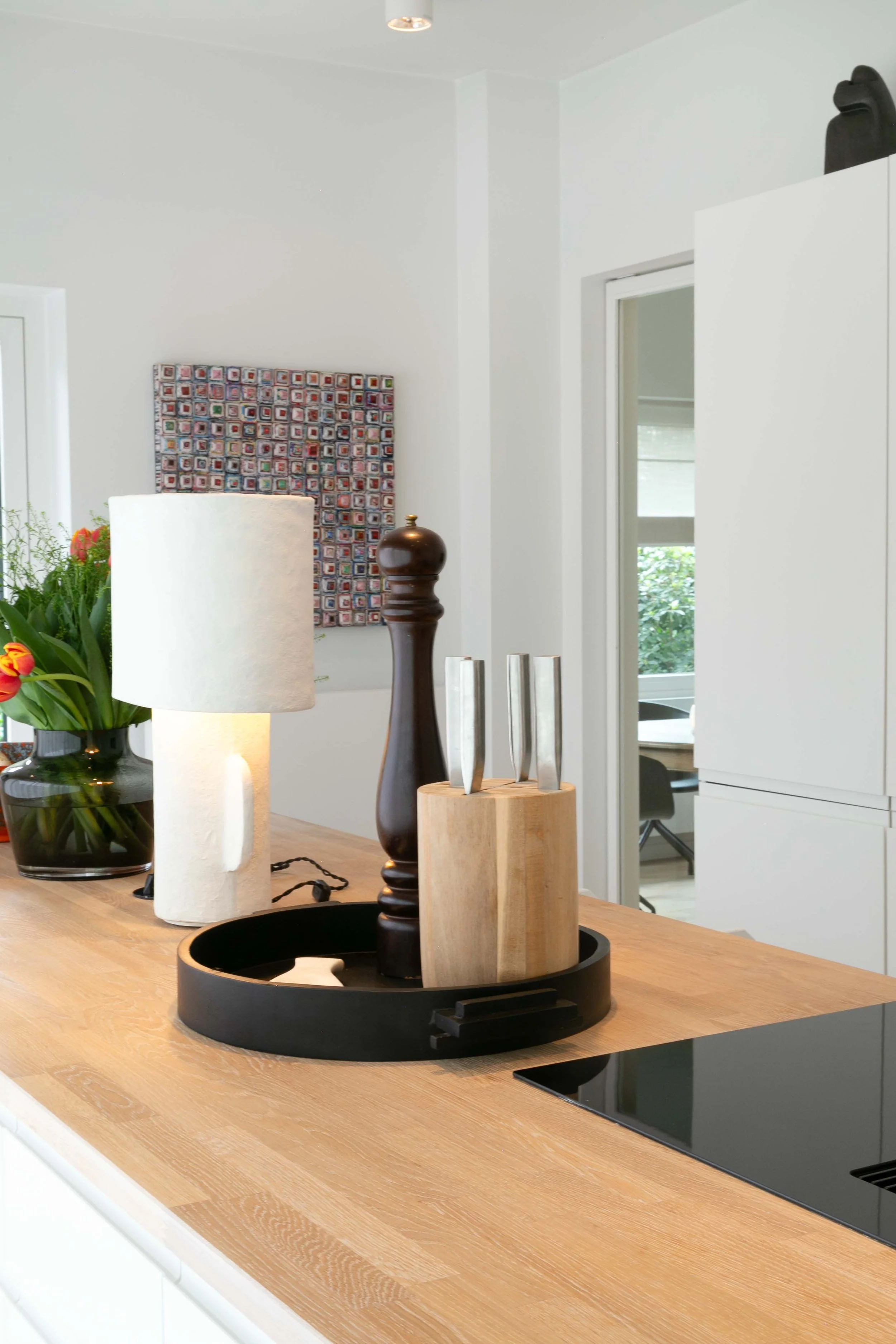 A wooden kitchen countertop with a black tray holding a variety of candles and décor pieces, a white textured paper towel roll, and a black vase with orange tulips, with a wall-mounted square mosaic art piece, a white lamp, and some greenery in the b