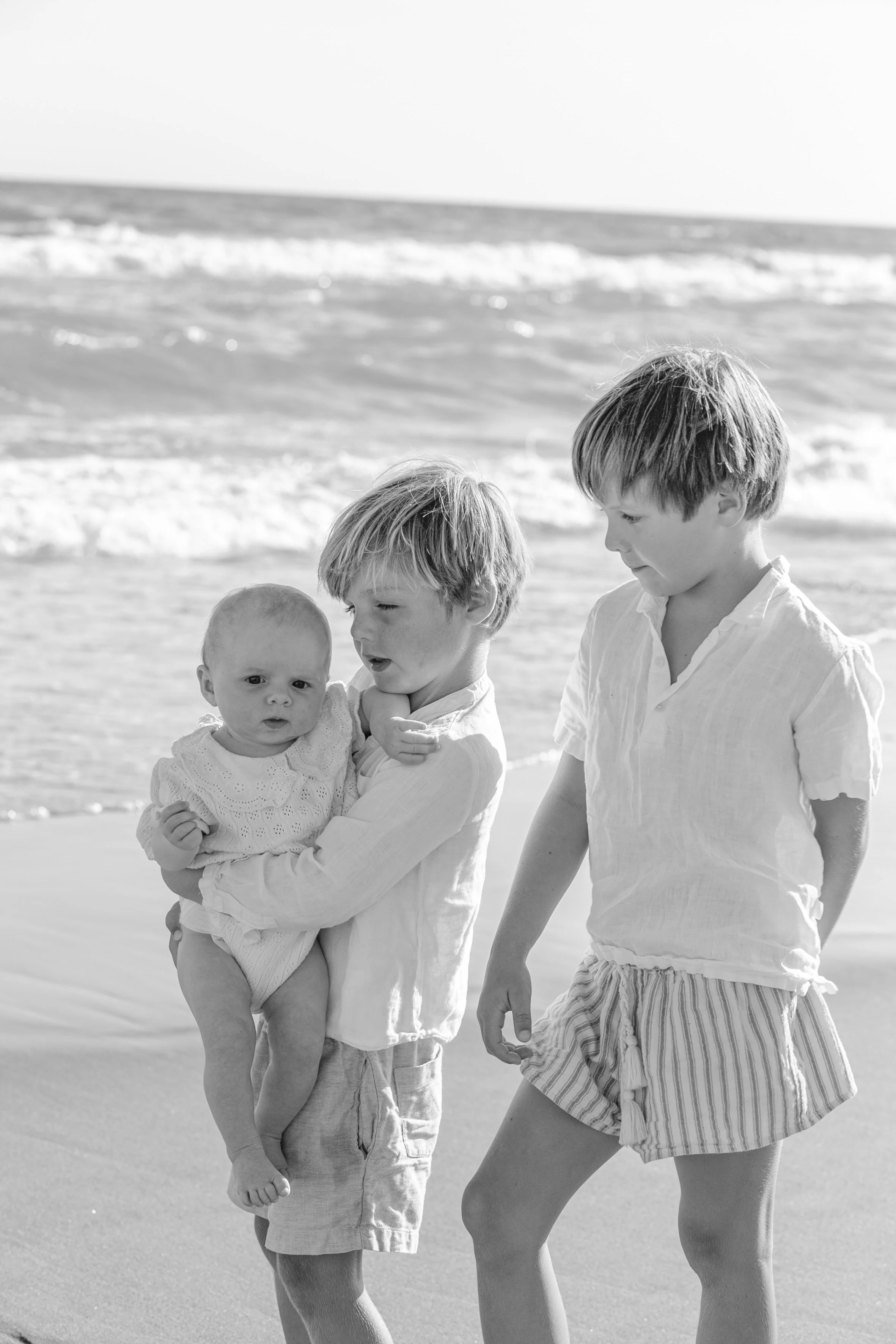 Three children on a beach in black and white, with the ocean in the background. The youngest child is being held by the middle child, who is looking at the oldest child. All three children are dressed in light-colored clothing.