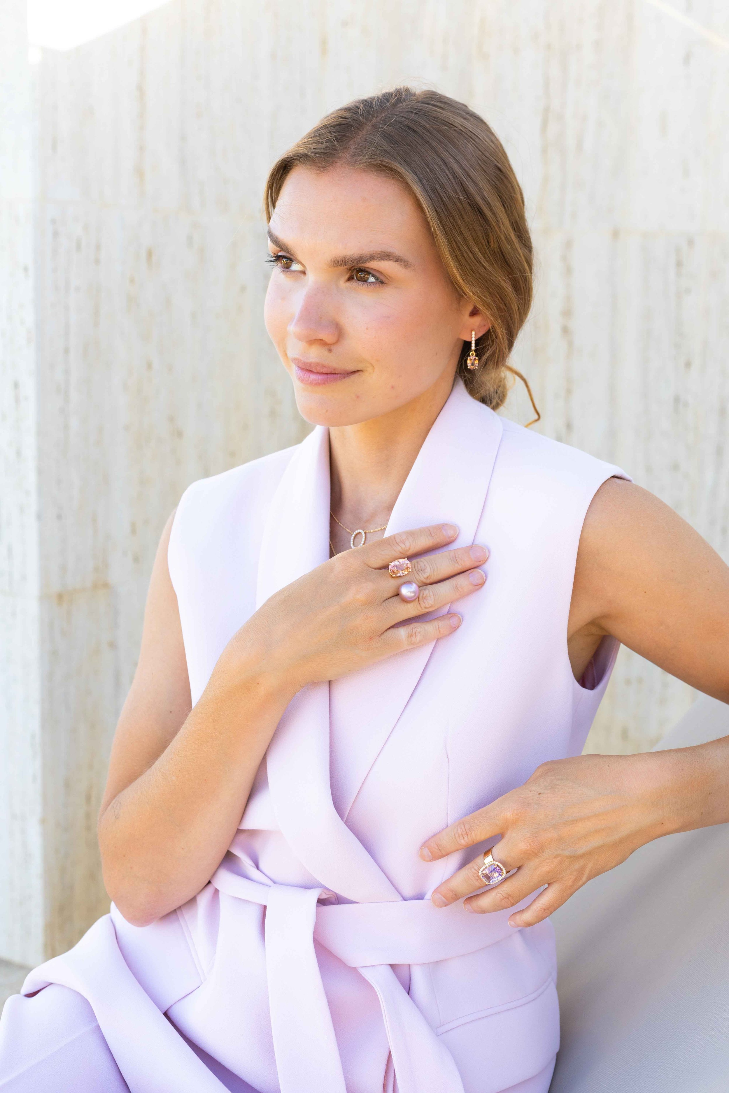 A woman with light brown hair styled in a braid, wearing a pale pink sleeveless dress and jewelry with pink and pearl accents, standing against a light-colored textured wall, looking thoughtfully to her left.