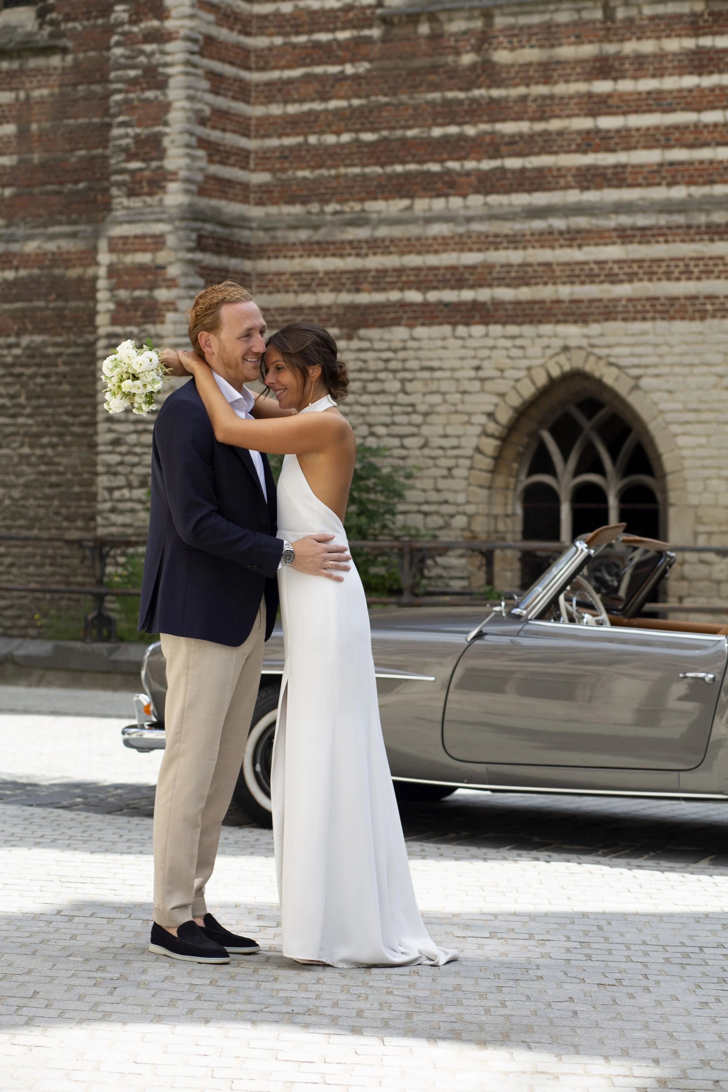 A man and woman dressed in wedding attire are smiling and embracing each other outdoors in front of a vintage car and a brick building. The man is wearing a navy blazer, beige pants, and black shoes. The woman is in a white wedding dress holding a bo
