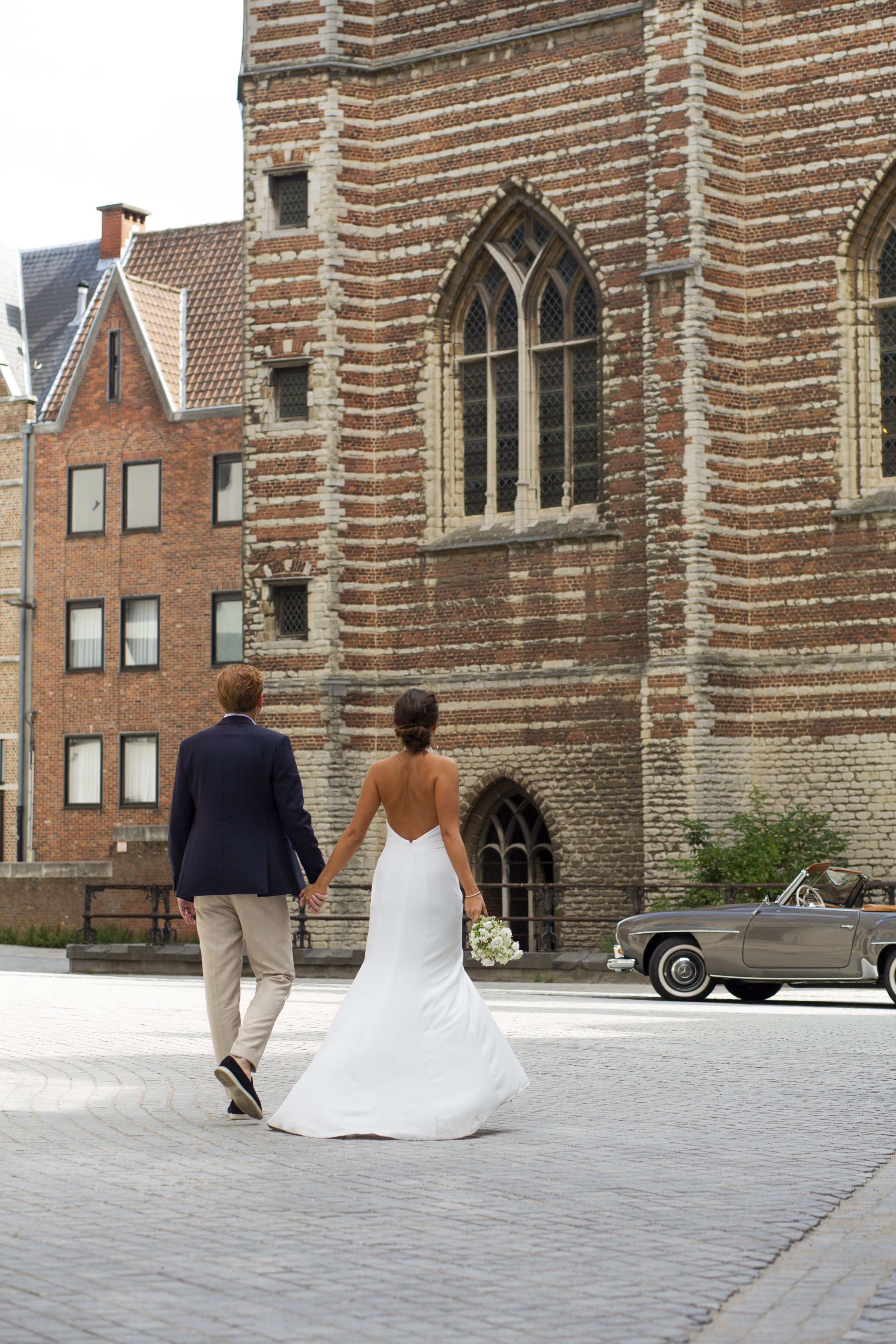 A newlywed couple holding hands and walking away from the camera in front of an old brick church, with a vintage silver car parked nearby.