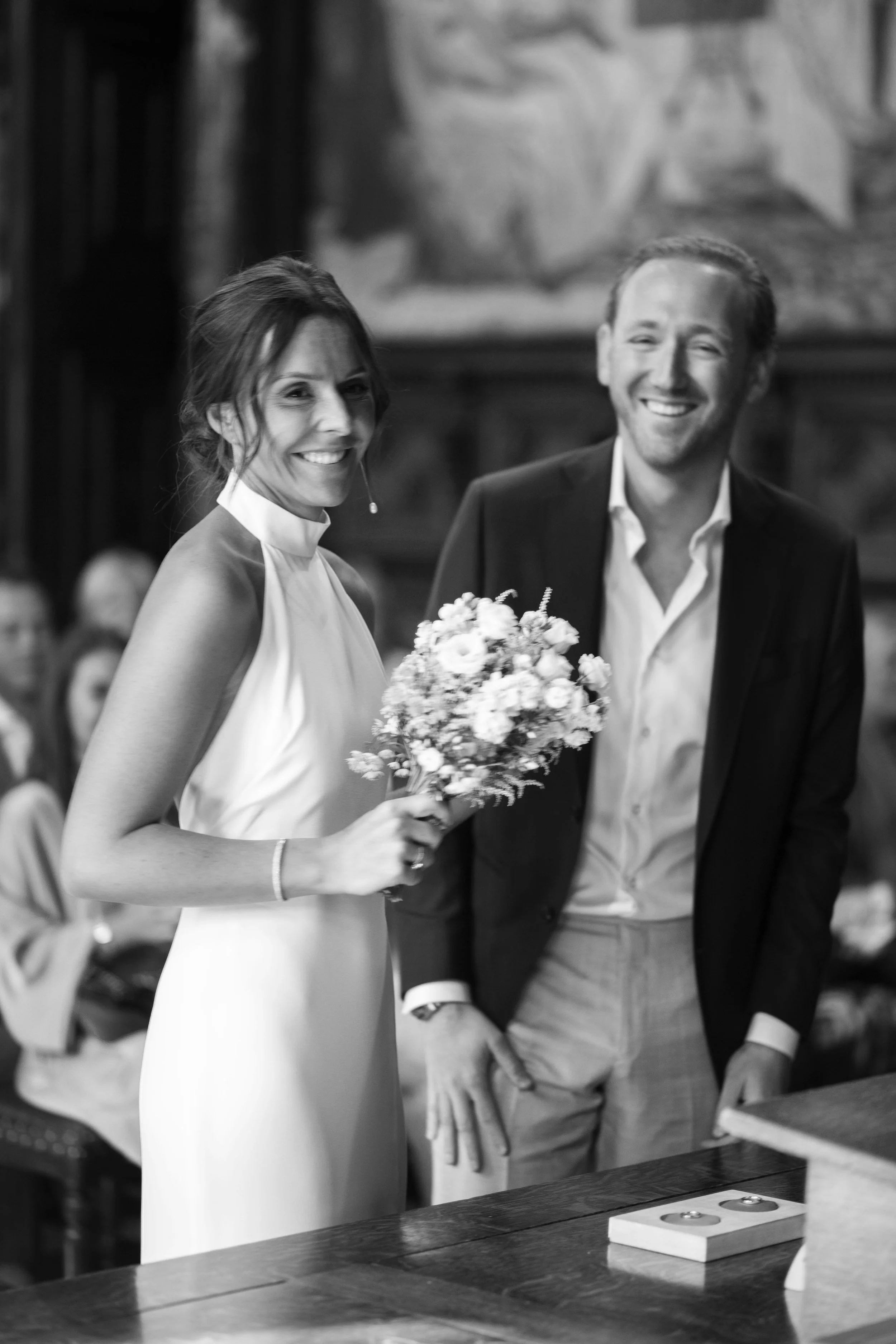 A black and white photo of a smiling woman in a sleeveless dress holding a bouquet and a man in a suit jacket and open-collared shirt standing next to her, during a wedding ceremony indoors.