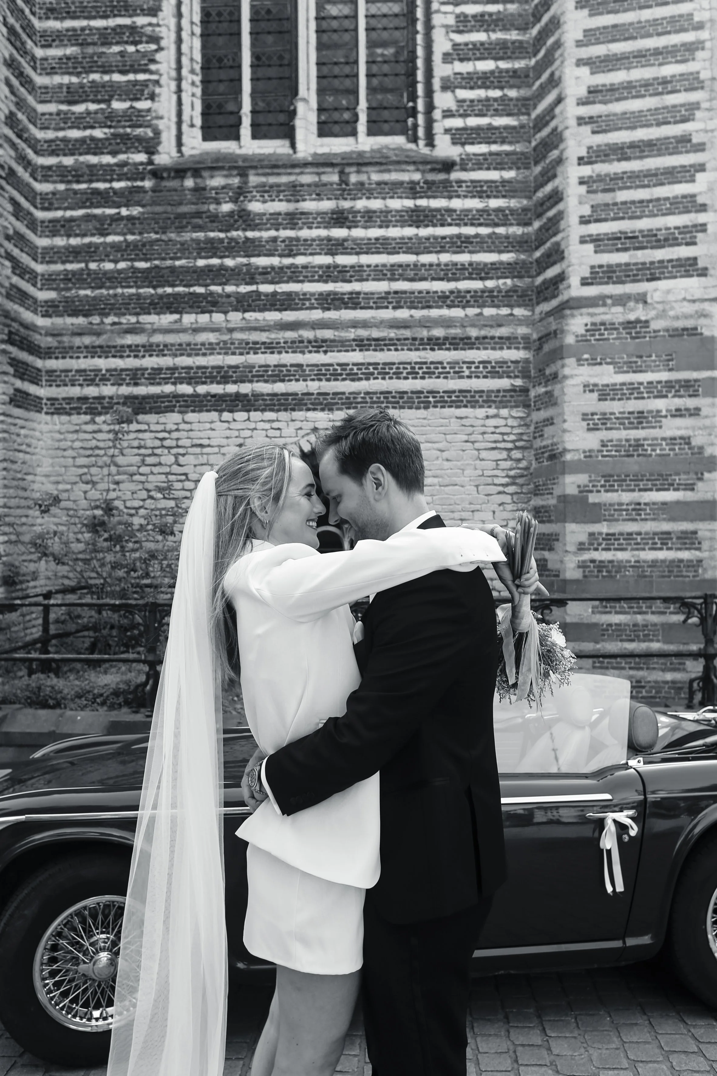 A bride and groom are sharing a close moment, smiling with foreheads touching, in front of a vintage car and a brick building.