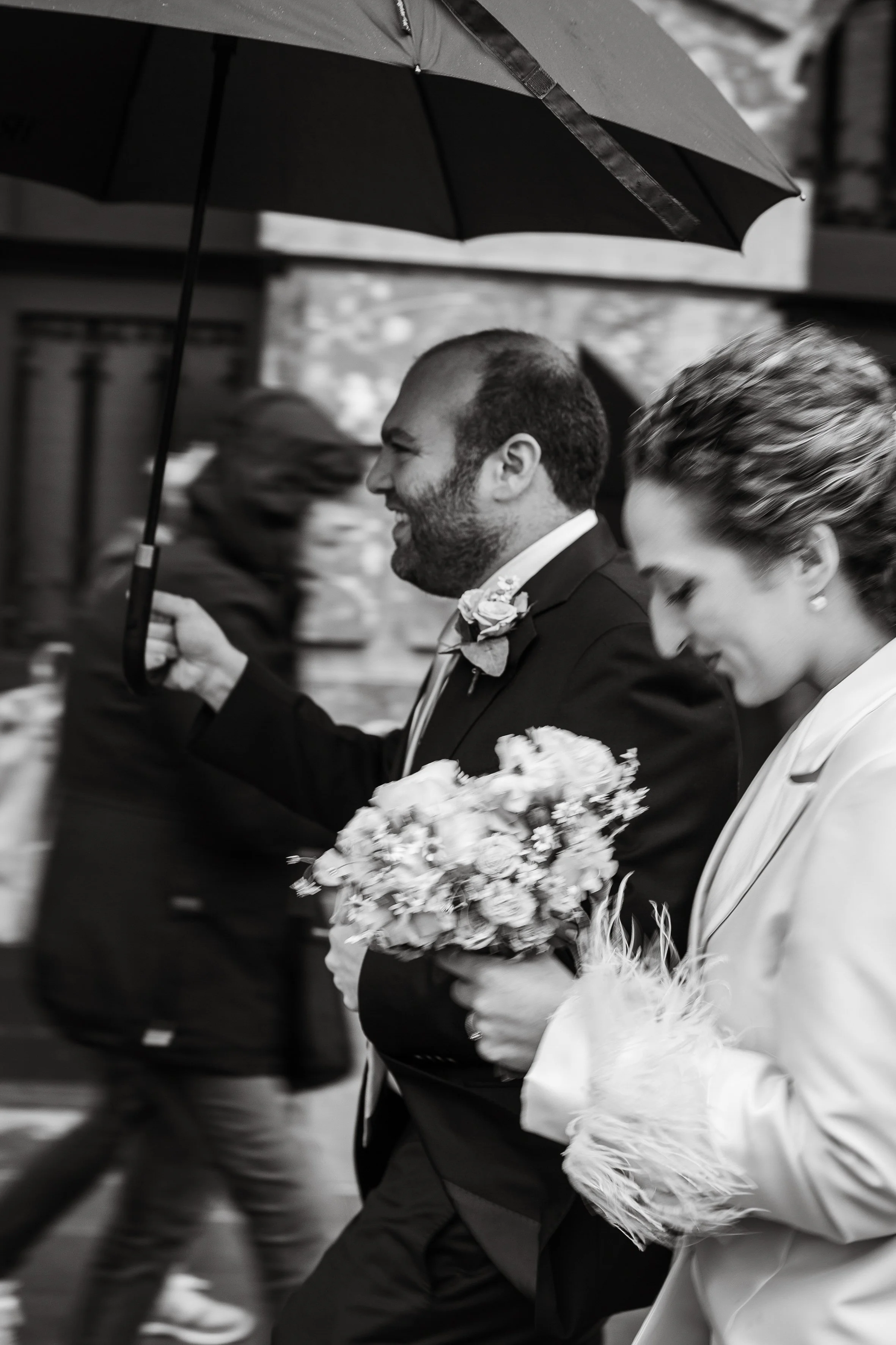 Black and white photo of a man and woman in formal attire walking during a rain shower, holding umbrellas and flowers.