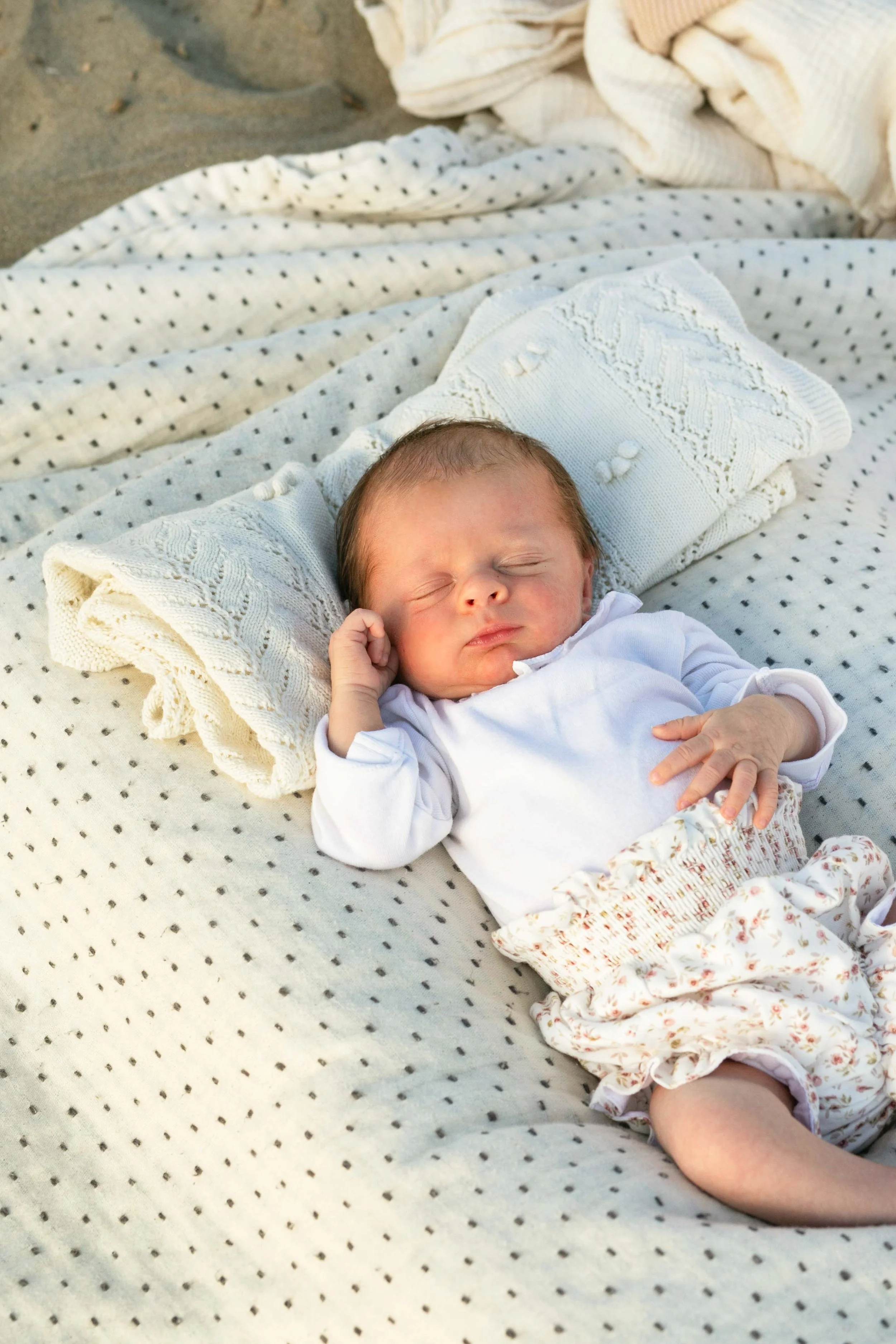 A sleeping baby lying on a blanket with polka dots, surrounded by textured white and floral blankets, on a sandy surface.