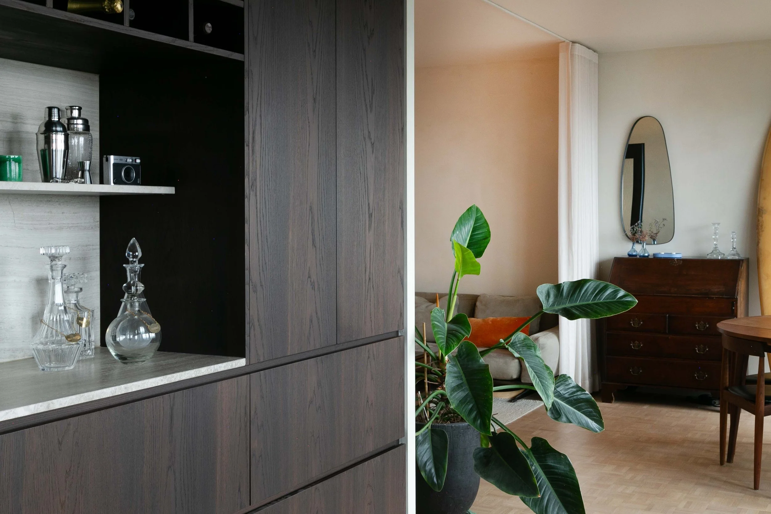 A living room with a dark wooden cabinet, glassware, and a potted plant in the foreground. In the background, there is a beige wall with a mirror, a wooden dresser, and a sofa.