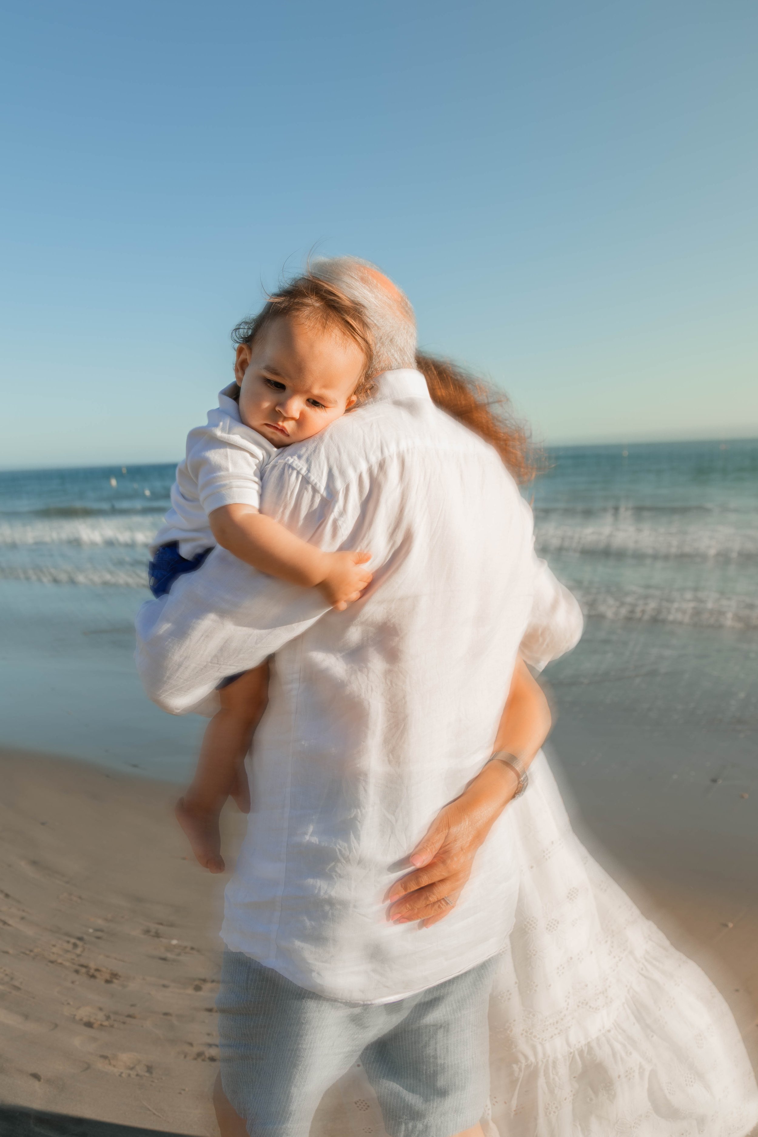 A person holding a young child at the beach with the ocean in the background. The child looks sad or pensive, and the person is wearing a white shirt and grey skirt or shorts. The sky is clear and blue.