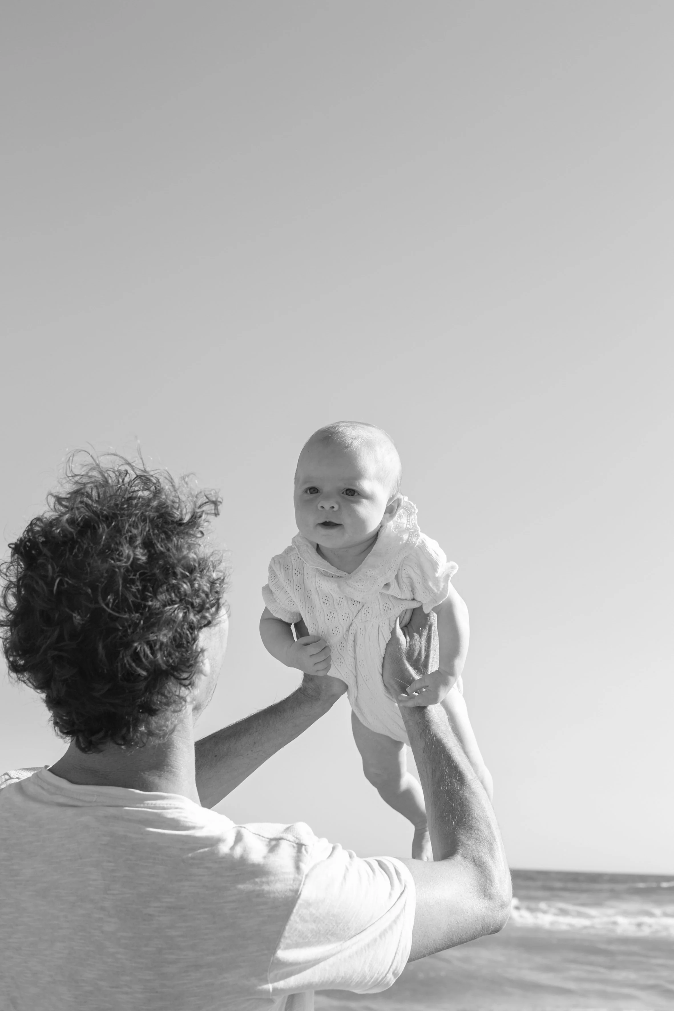 A man with curly hair holding a baby girl dressed in a white dress at the beach, with the ocean and sky in the background.