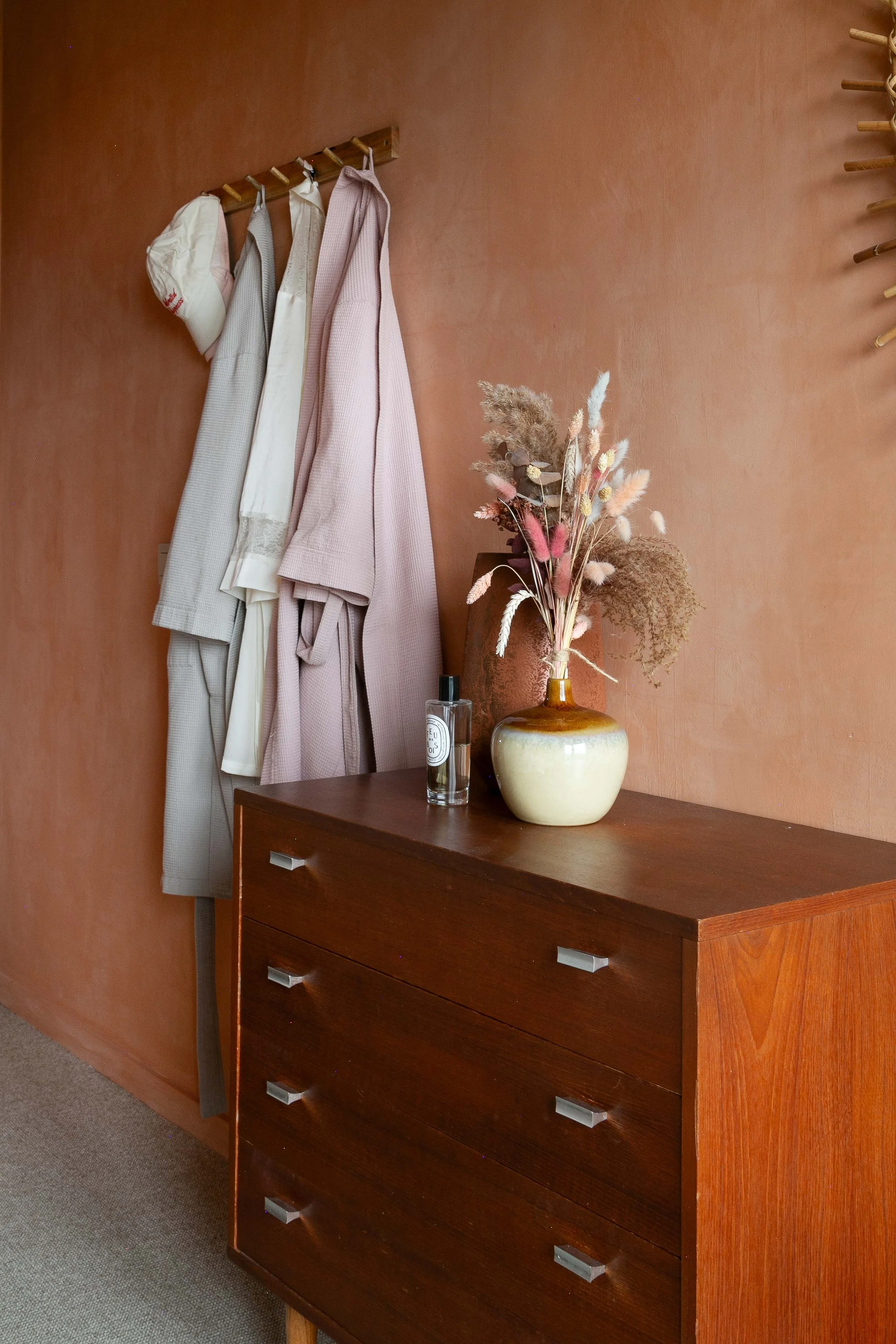 A wooden dresser with four drawers, next to a brown wall. On top of the dresser, there is a large beige and brown ceramic vase with dried flowers, a clear glass spray bottle, and hanging on the wall are bathrobes and a towel.