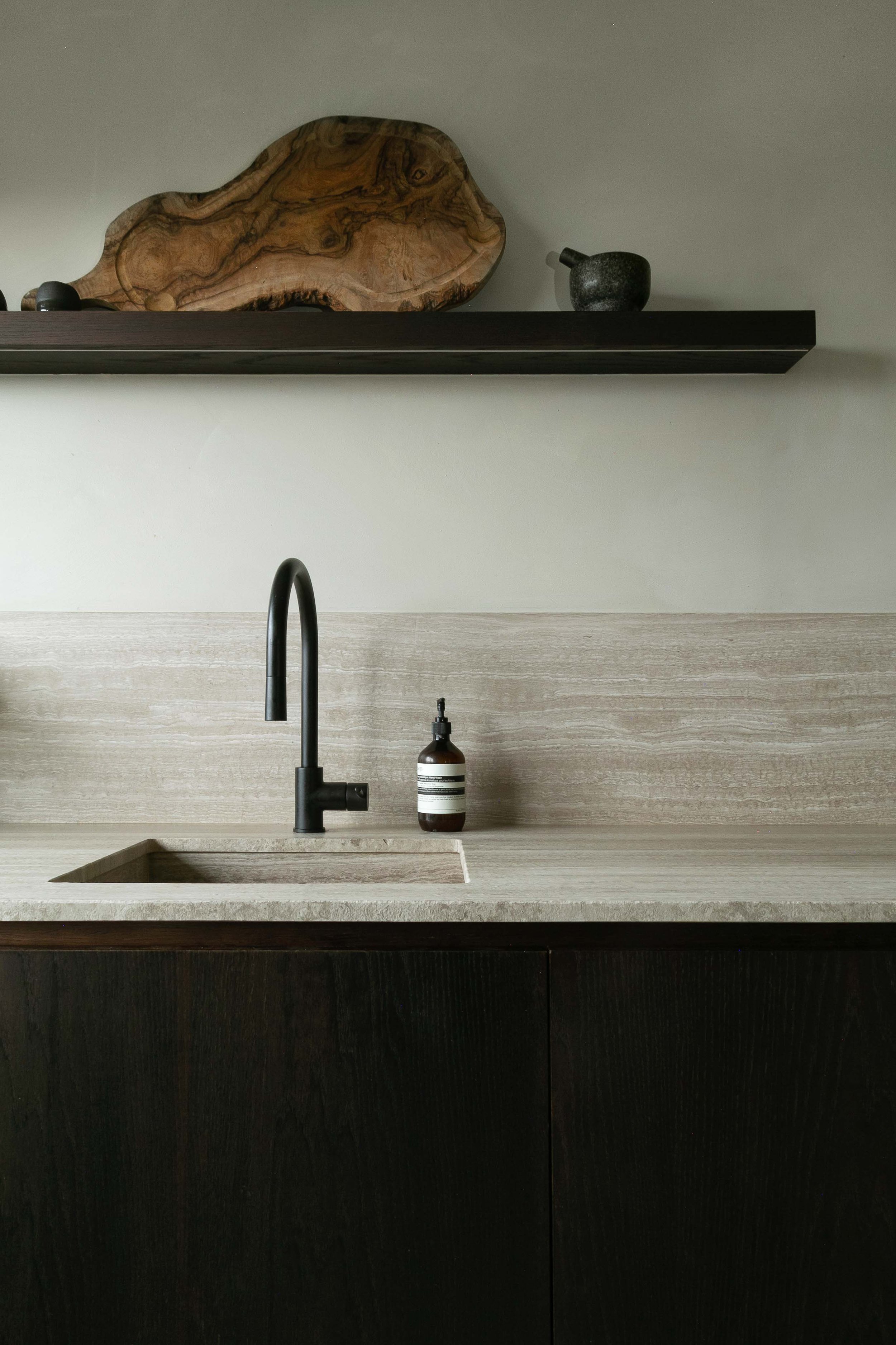 A modern kitchen with a black faucet, a beige countertop with an integrated sink, a dark wooden cabinet, and beige textured backsplash. A bottle of soap or lotion sits on the countertop. A floating wooden shelf with decorative items, including a larg