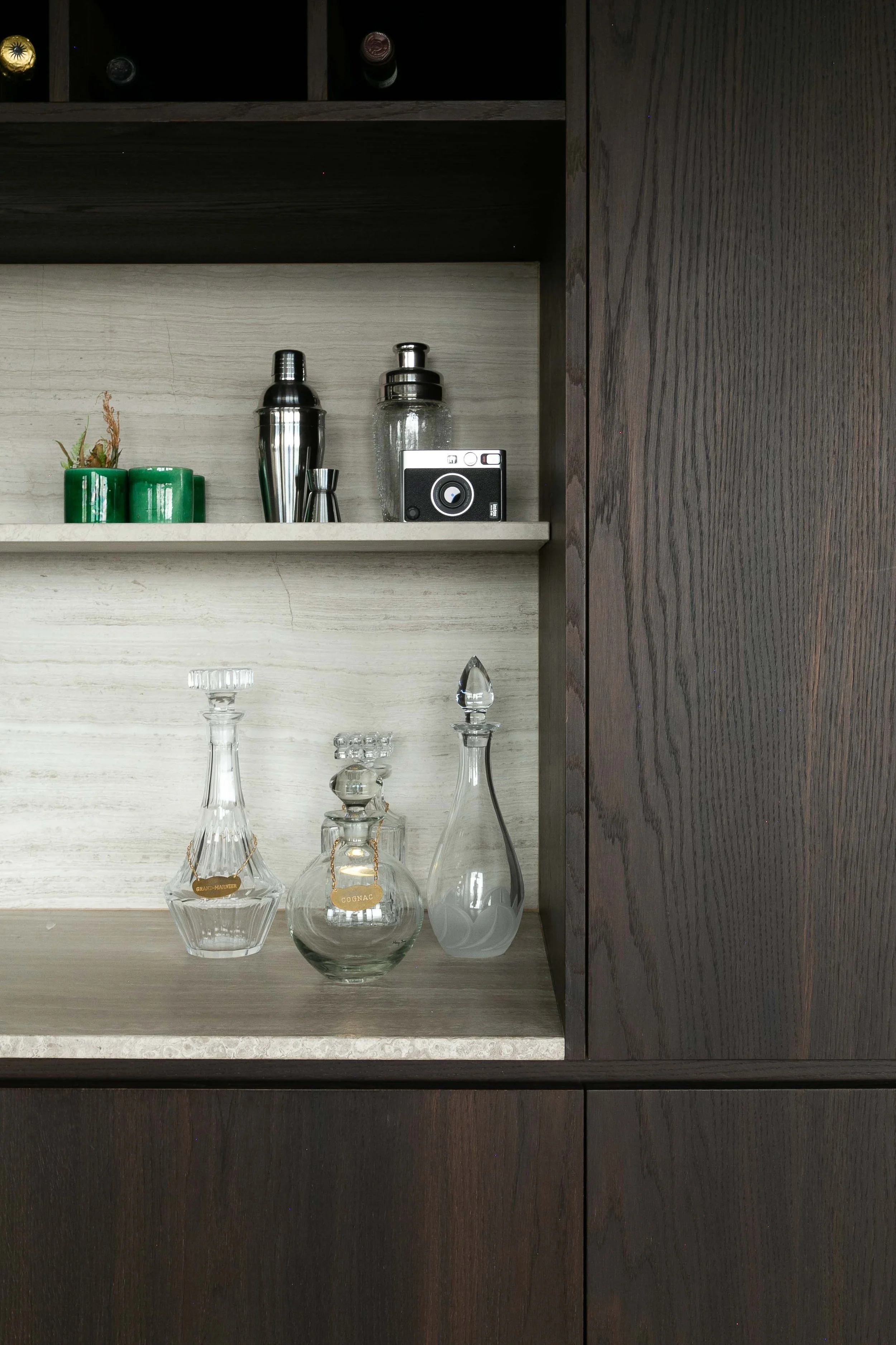 A wooden display cabinet contains various glassware, including decanters and glass bottles. On the upper shelf, there are small potted plants and bar tools, alongside a small camera. The cabinet has a dark wood frame and a light wood back panel.
