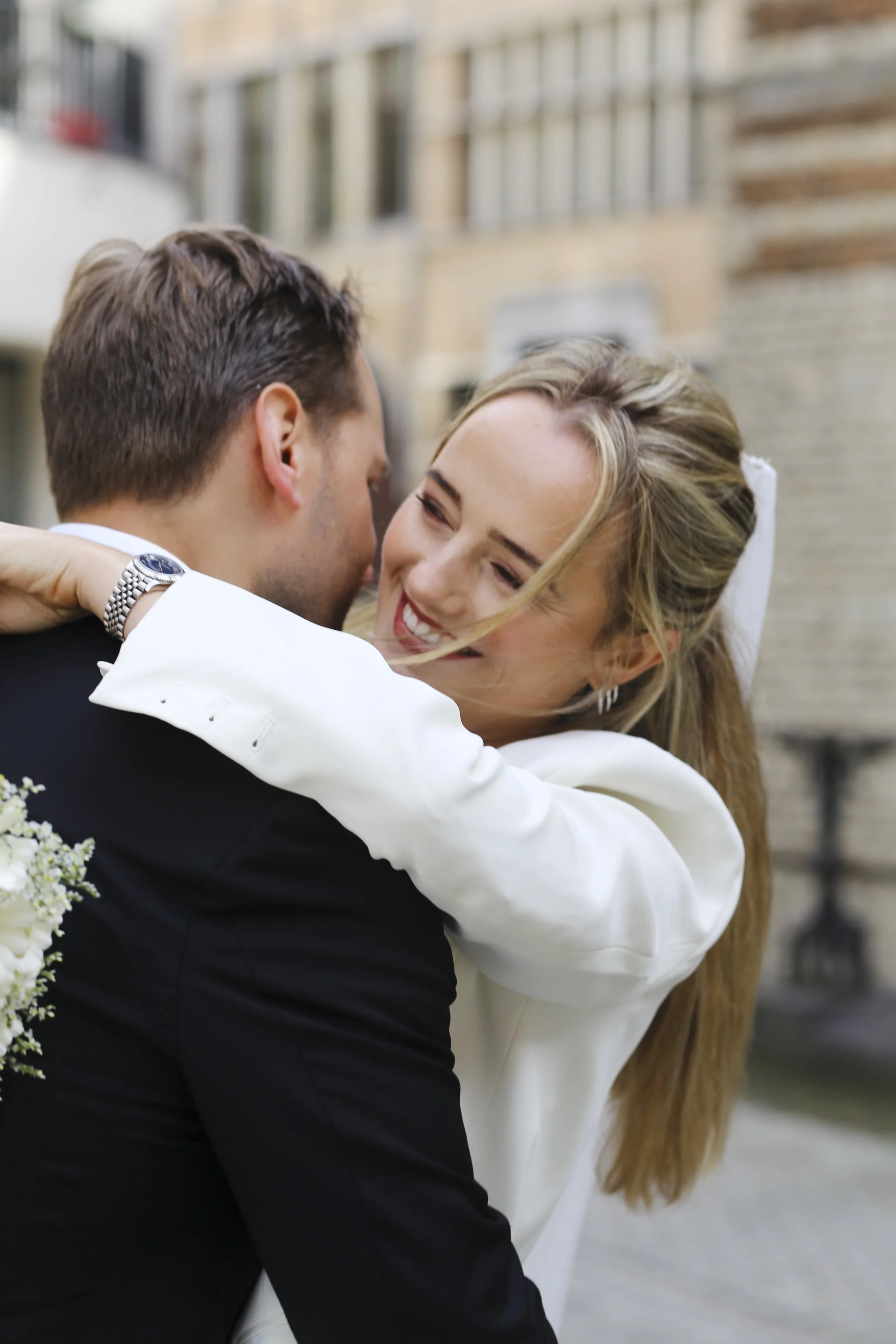 A bride and groom embrace each other and smile in an outdoor setting with a brick building in the background.