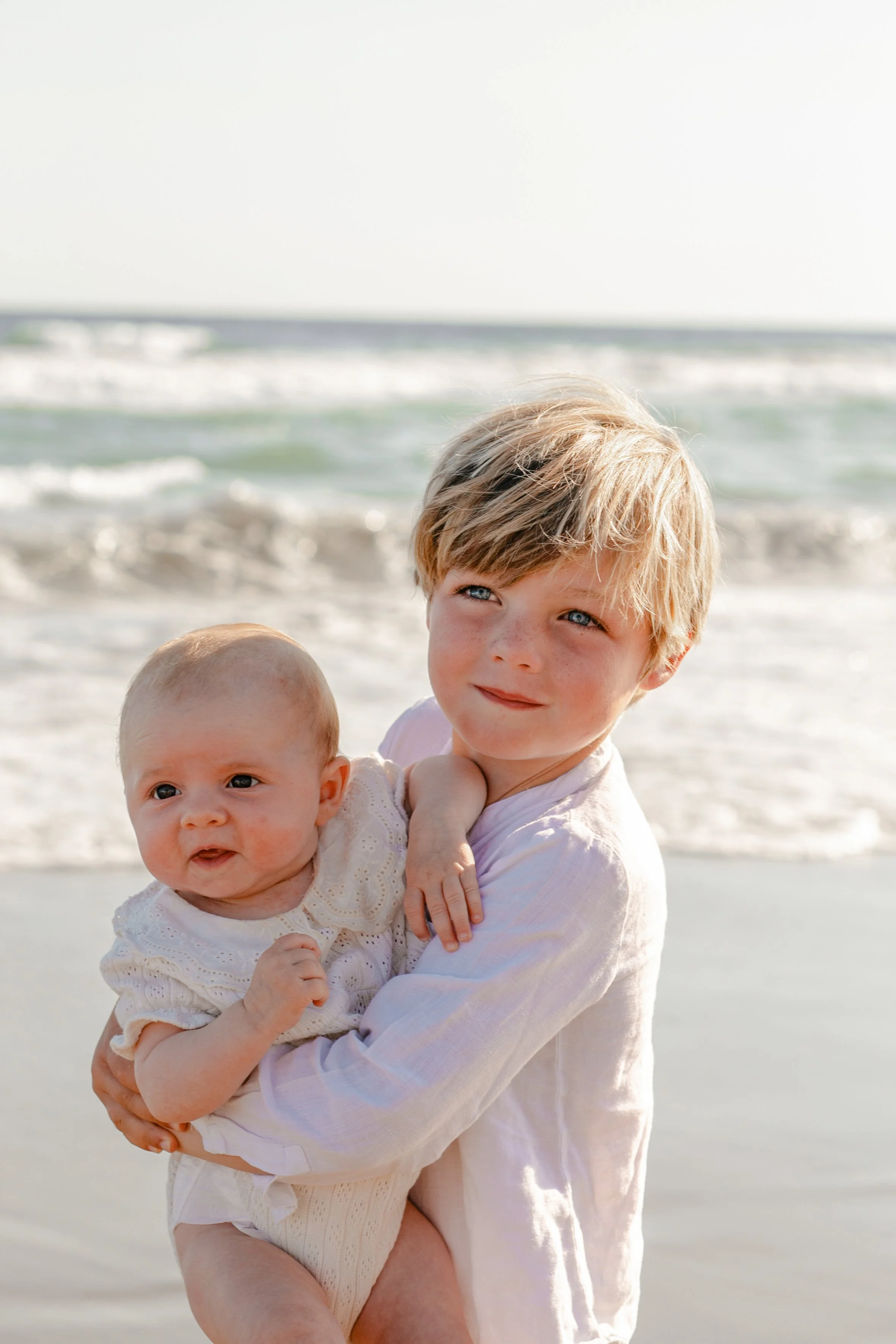 Two children, a young girl with blonde hair and a baby, are on a beach with waves in the background. The girl is holding the baby and both are looking at the camera.