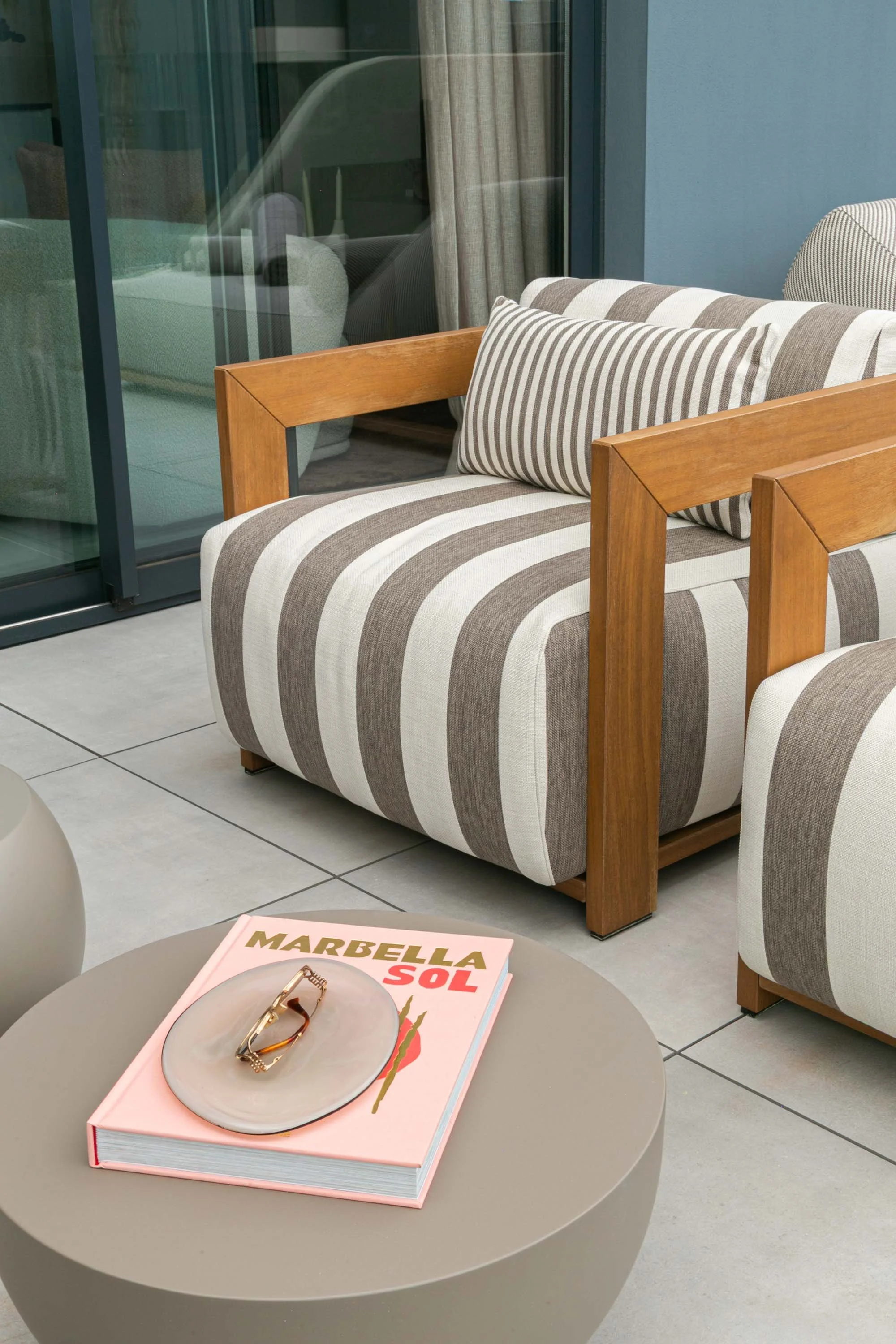 A pink hardcover book titled 'Marbella Sol' resting on a round beige table, with a pair of sunglasses on top of the book, in an indoor setting with striped armchairs and a glass door.