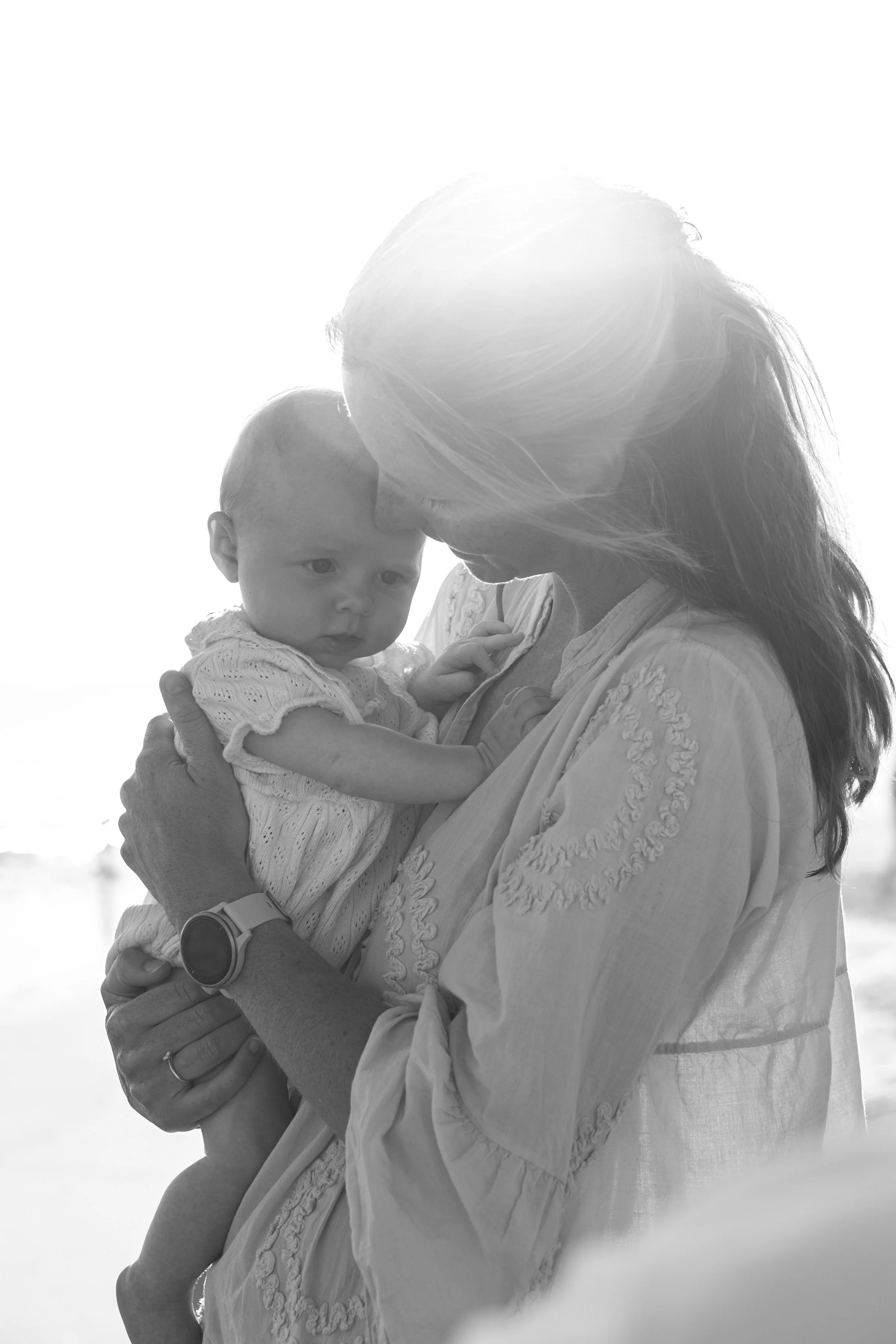 A woman with long hair holding a baby on her shoulder outdoors, backlit by sunlight.